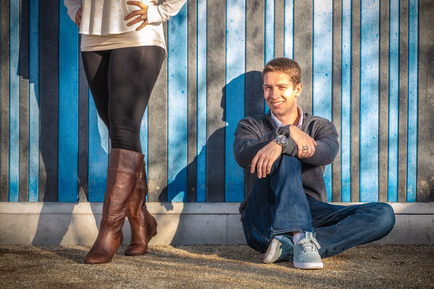 A seated person in blue leaning against a striped wall while another person in brown boots stands nearby.