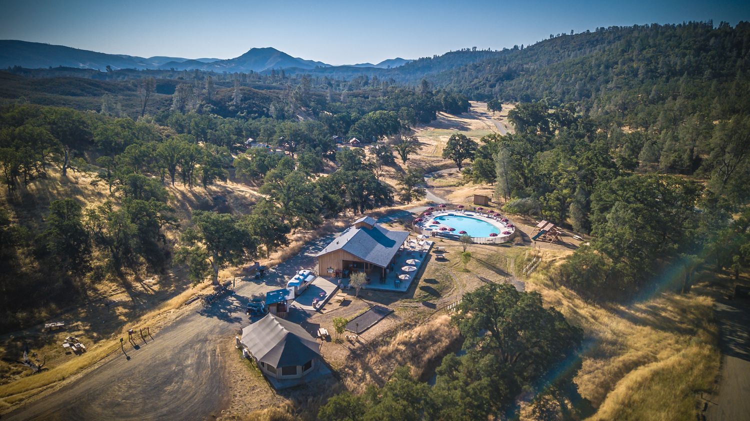 Aerial view of a luxury resort nestled in wooded mountains with swimming pool and modern buildings.