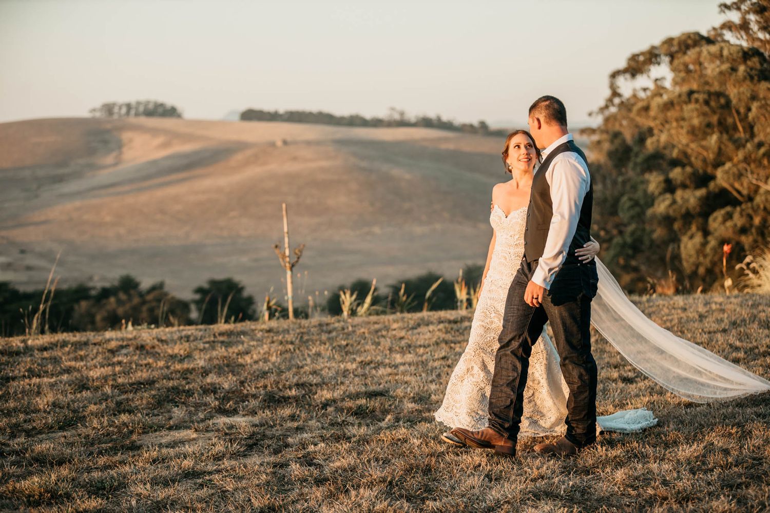 Newlyweds standing together on a hilltop during golden hour with flowing veil and rolling hills in background.