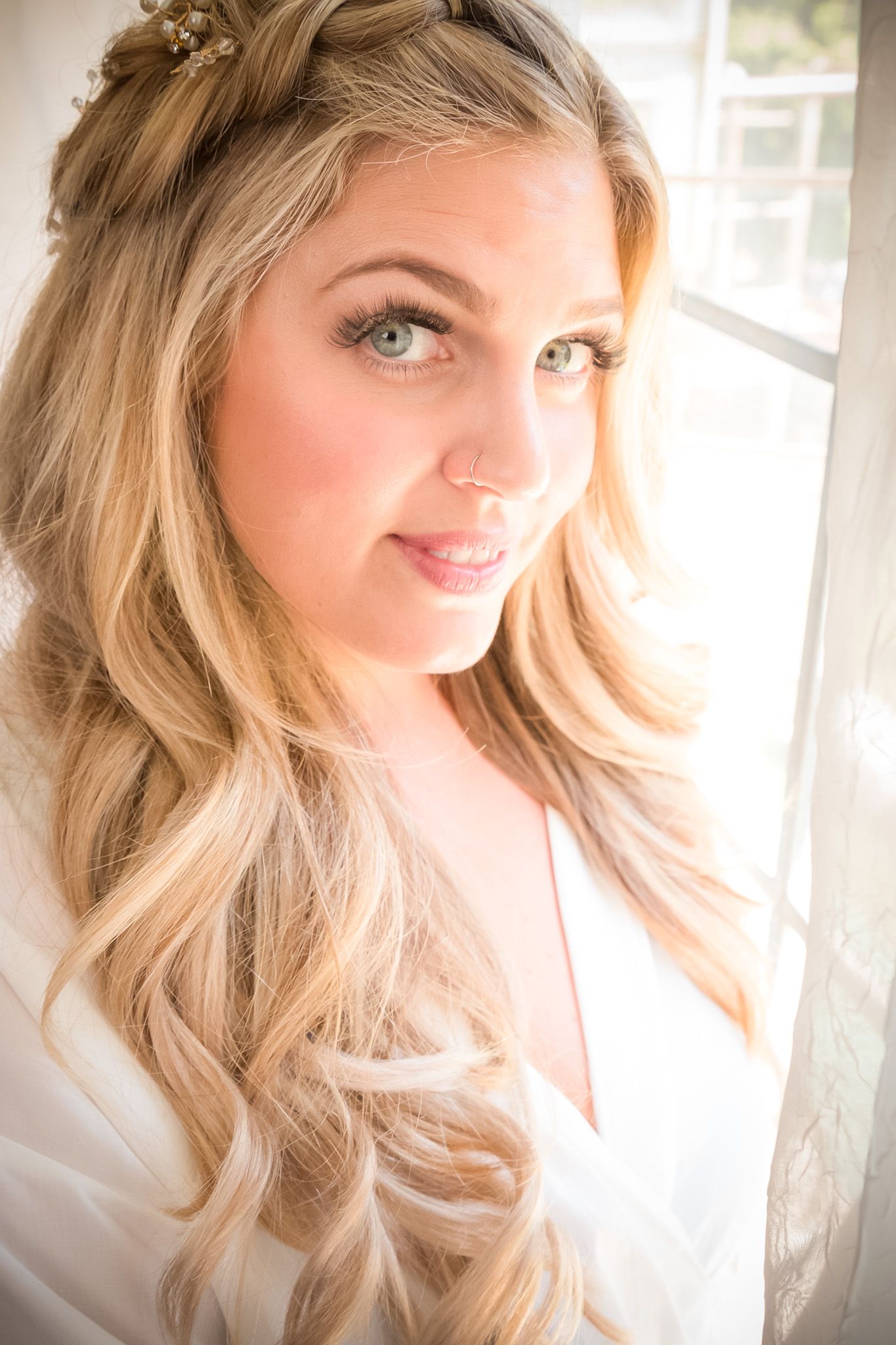 Soft portrait of a bride with flowing blonde hair and floral crown near window.