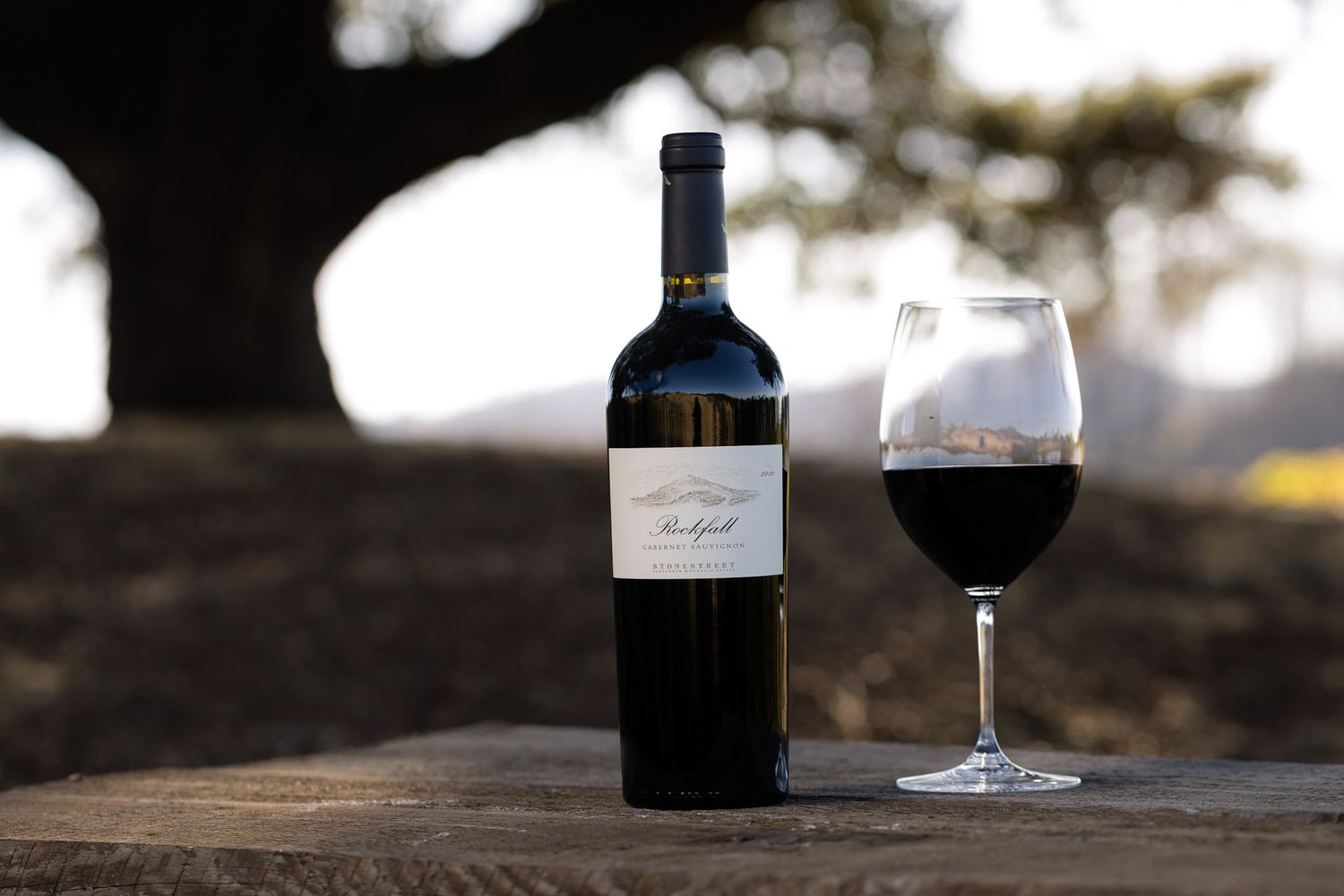 A bottle of red wine and filled wine glass on a rustic wooden surface at sunset.