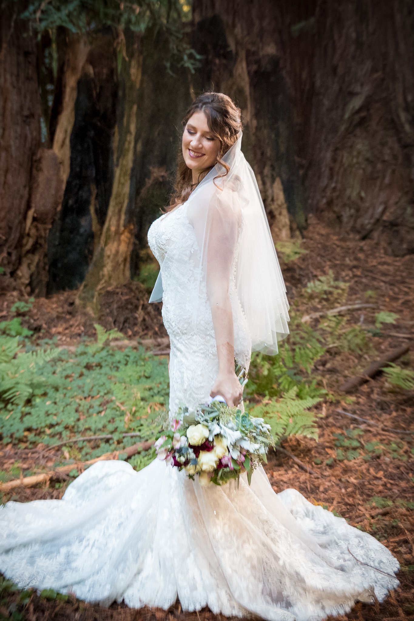A bride in a form-fitting lace gown with long veil poses with her bouquet against a backdrop of tall trees.