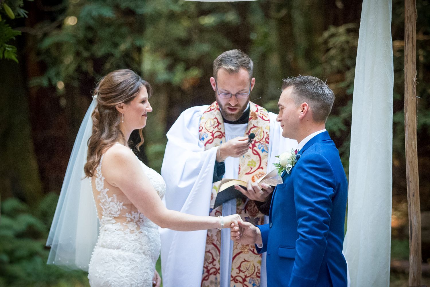 A wedding ceremony in a forest setting with bride, groom and officiant exchanging vows under white draping.
