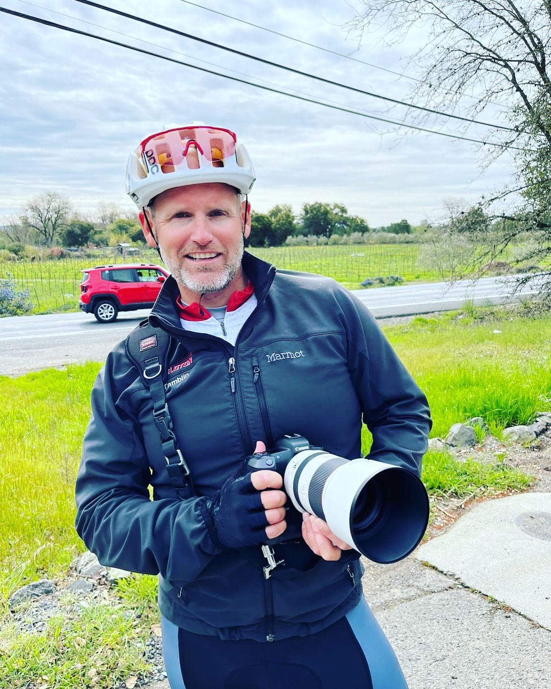 Photographer in cycling gear holding a large camera lens outdoors.