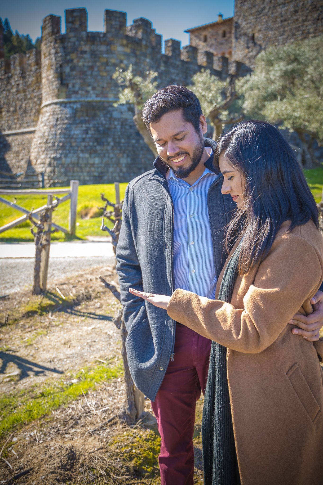 A couple looks at something together while standing in front of a stone castle wall on a sunny day.