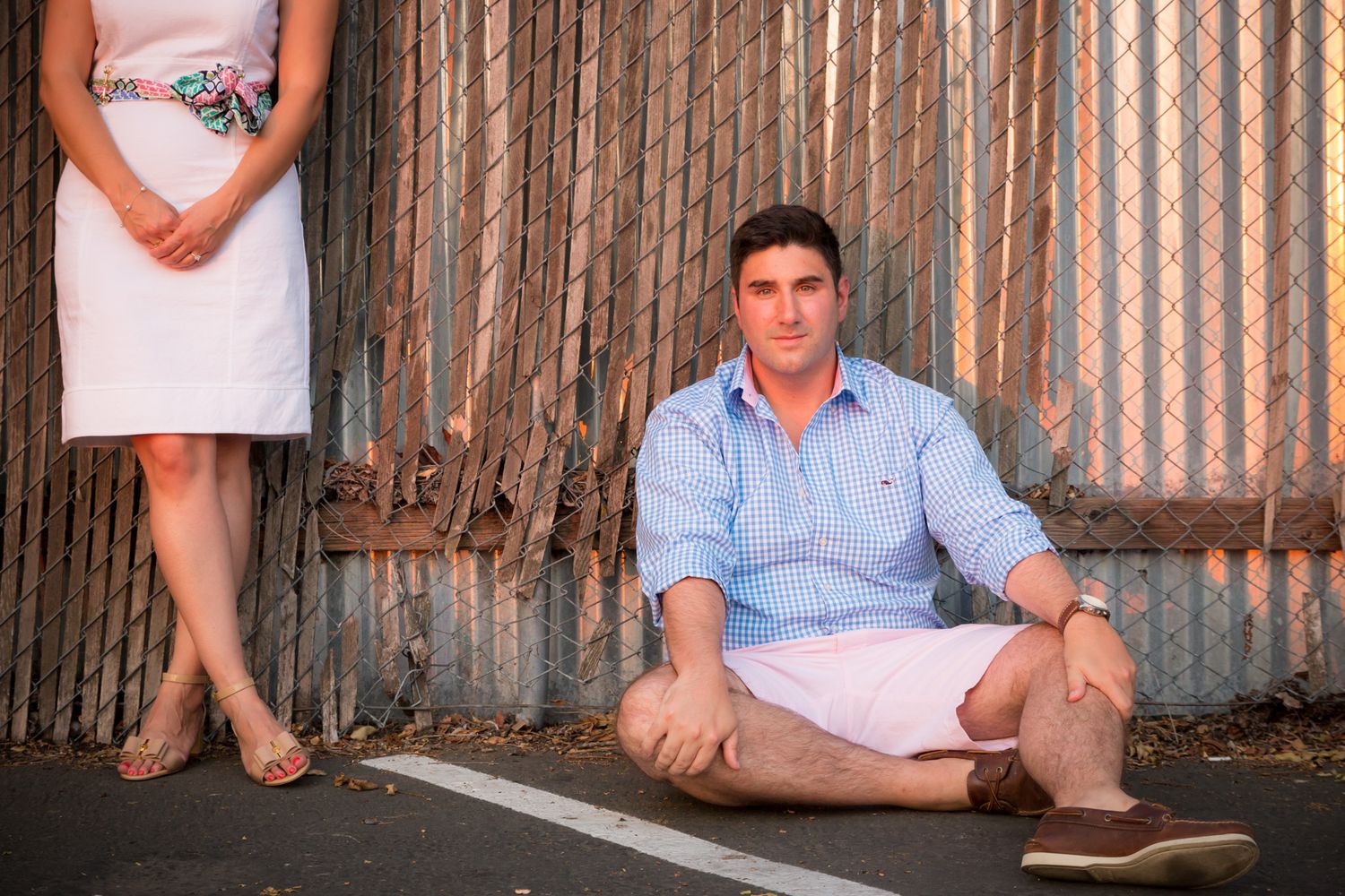 A person sits on the ground in a blue shirt and pink shorts while another stands nearby in a white dress.
