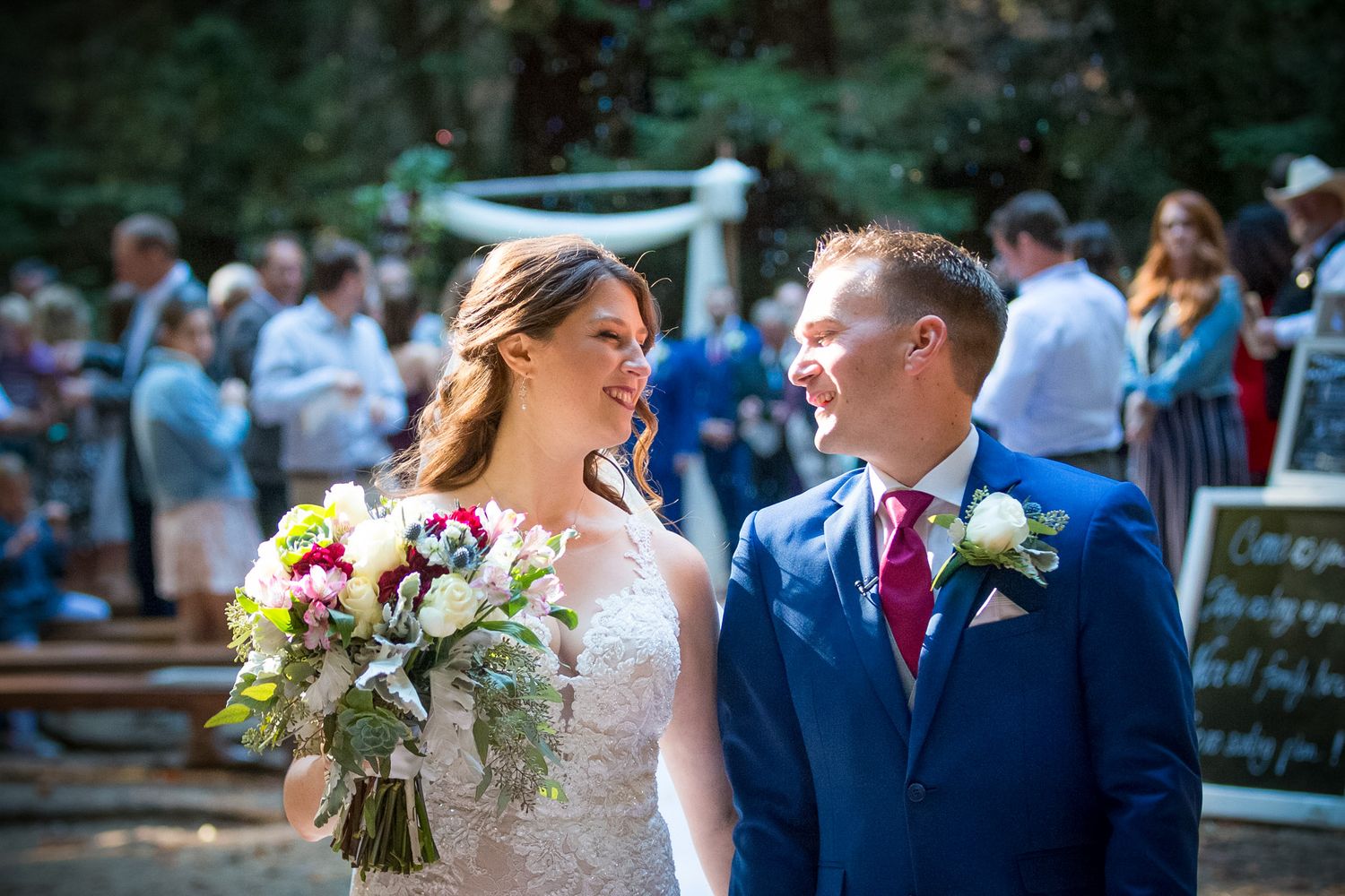 Bride in lace dress and groom in blue suit share a joyful moment during their forest wedding ceremony.