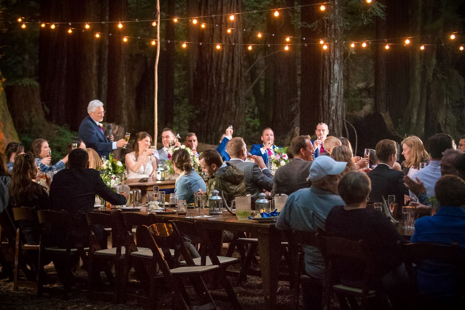 Outdoor wedding reception under string lights in a redwood forest with guests seated at long tables.