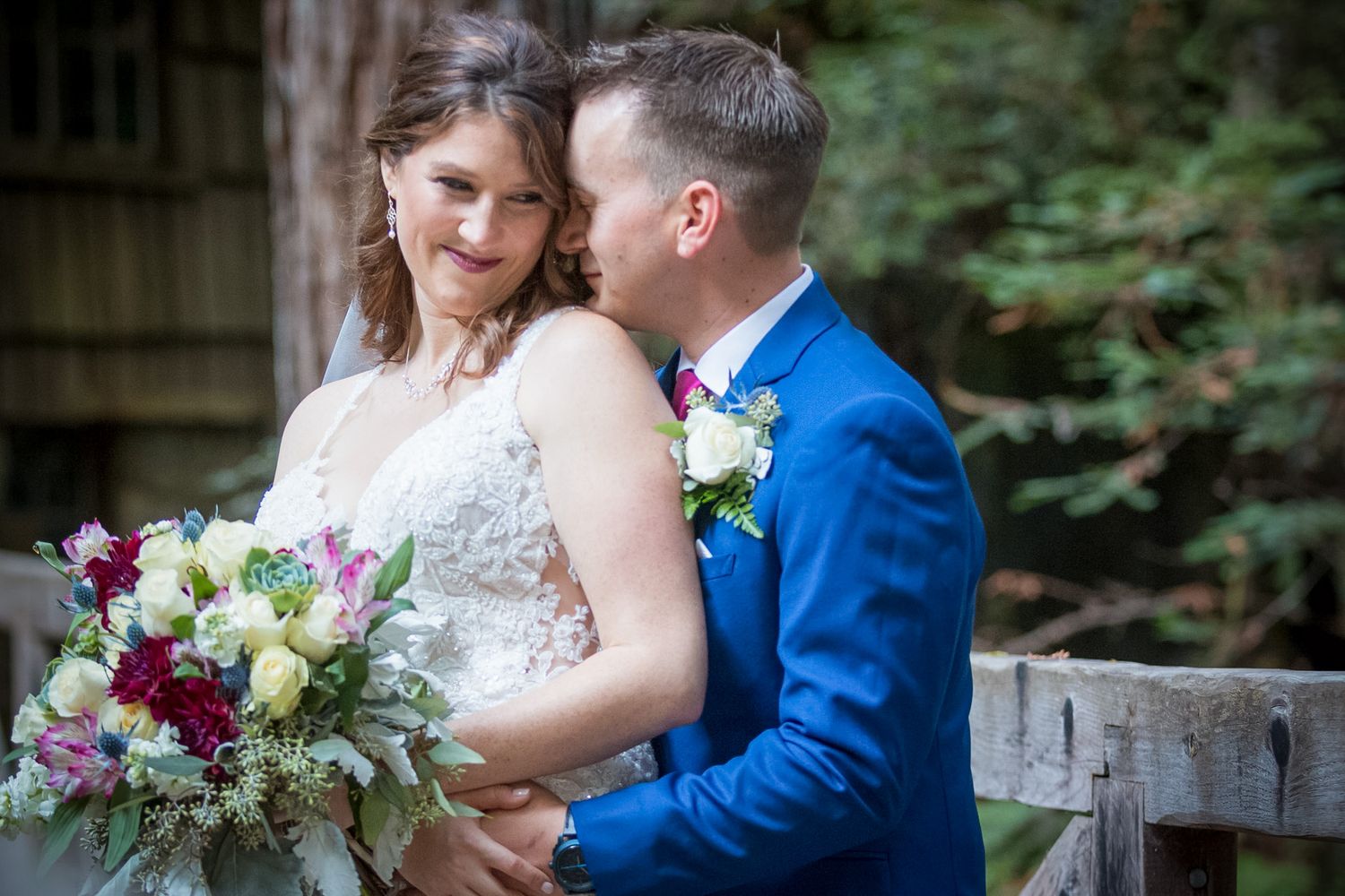Tender moment between newlyweds with burgundy and white bouquet against forest backdrop.