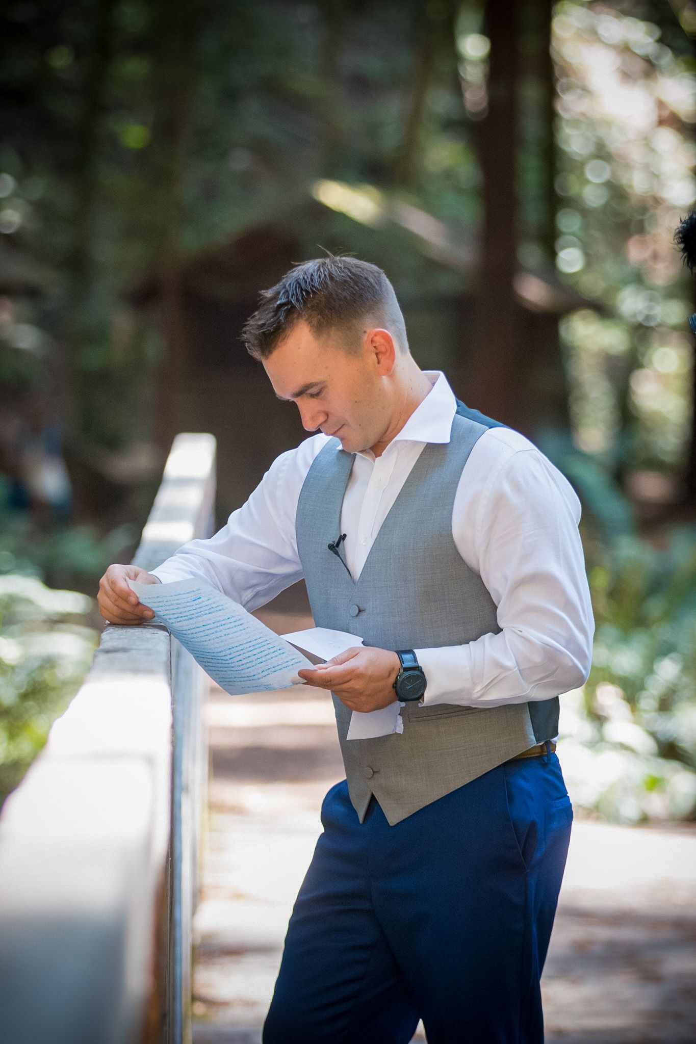 Groom in grey vest and navy pants reads document while standing on wooden walkway in forest.