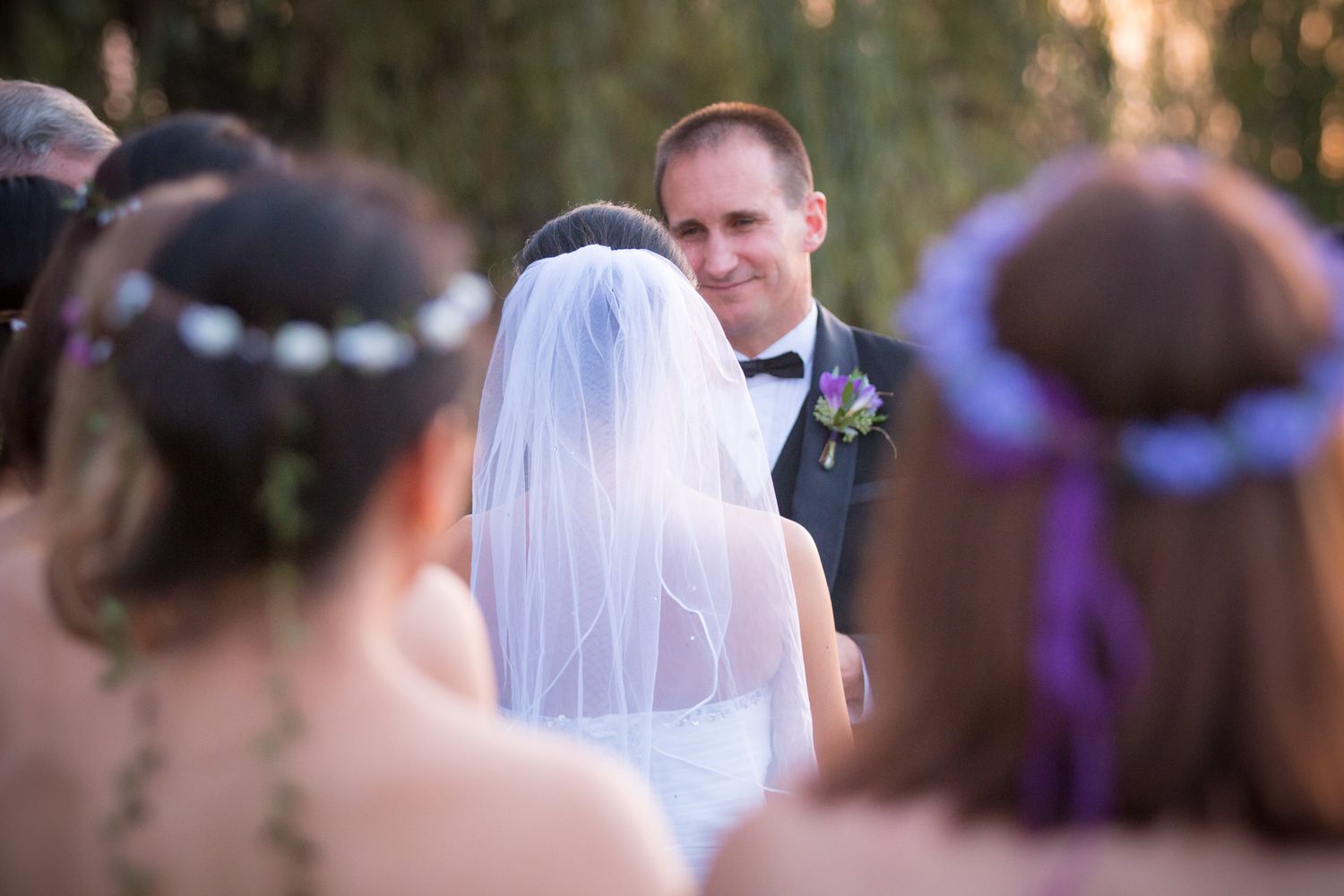 Wedding scene with bridesmaids wearing purple floral crowns watching as bride in white veil faces groom during ceremony.