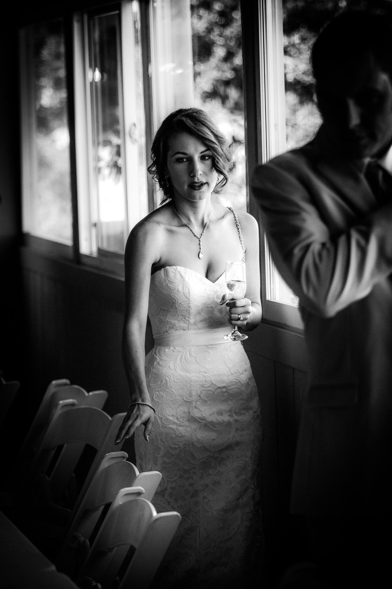 A black and white wedding photograph showing a bride in a strapless lace gown standing by a sunlit window.