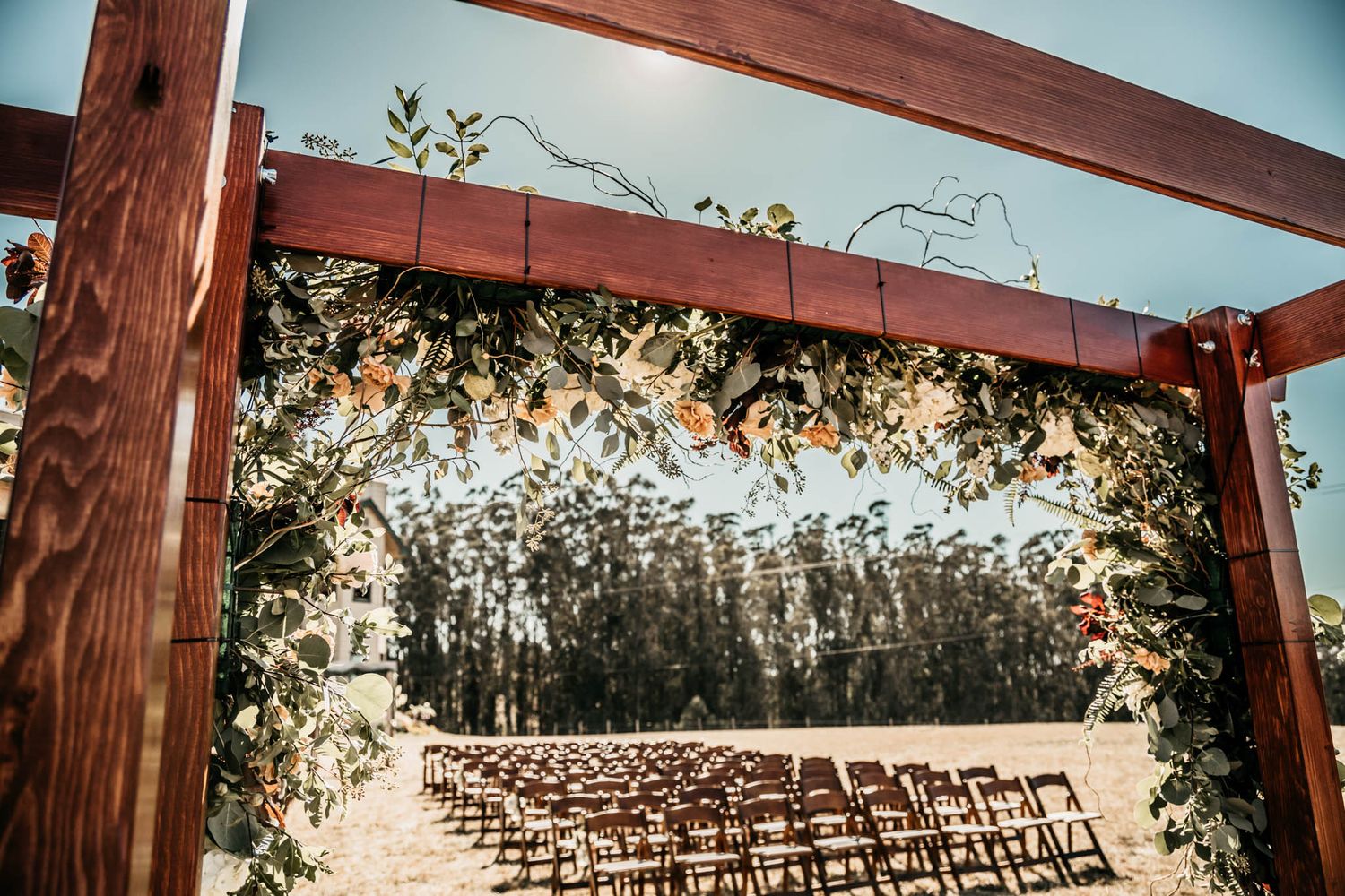 Wedding ceremony setup with wooden arch decorated in flowers and white chairs lined up outdoors.