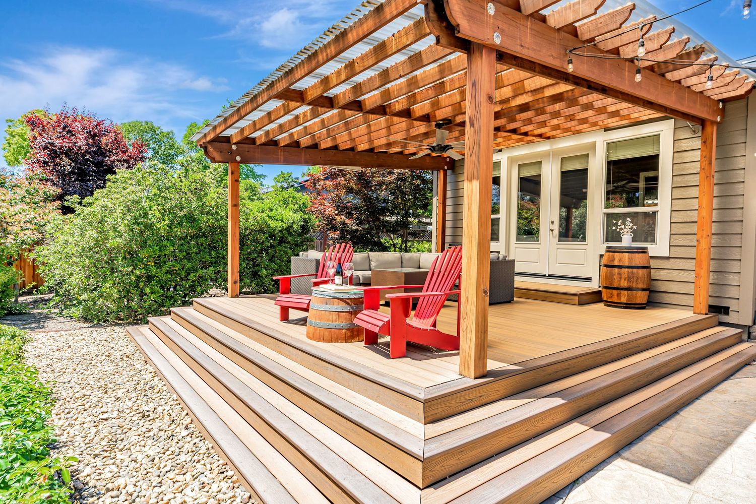 Modern wooden pergola deck with red Adirondack chairs and wine barrel accent in a landscaped backyard.