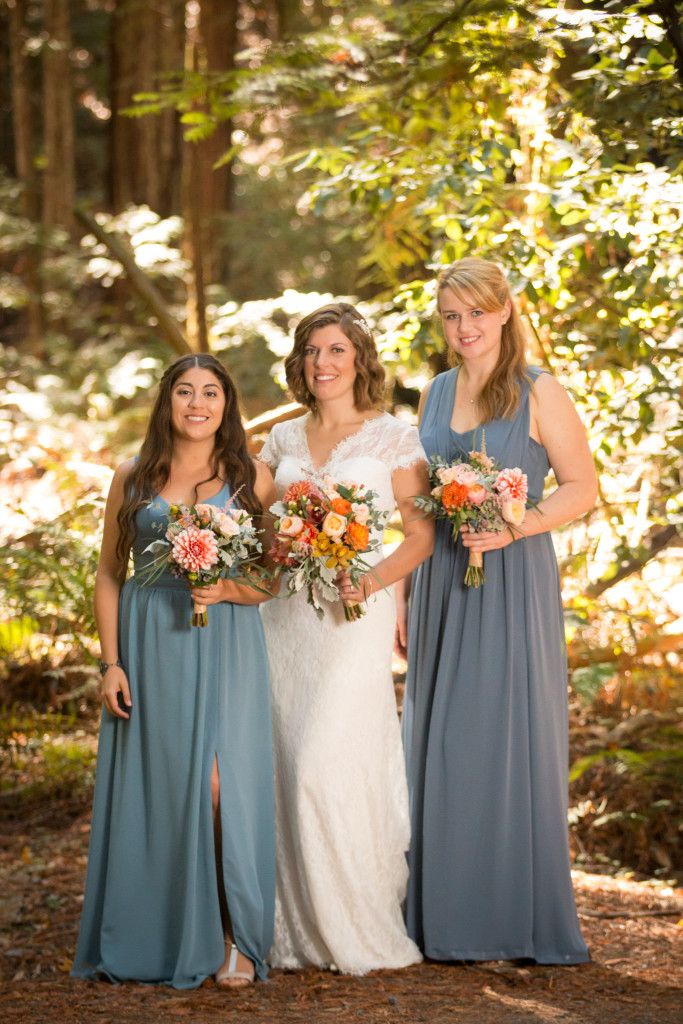 Three women pose in sunlit forest wearing white wedding gown and dusty blue bridesmaid dresses.