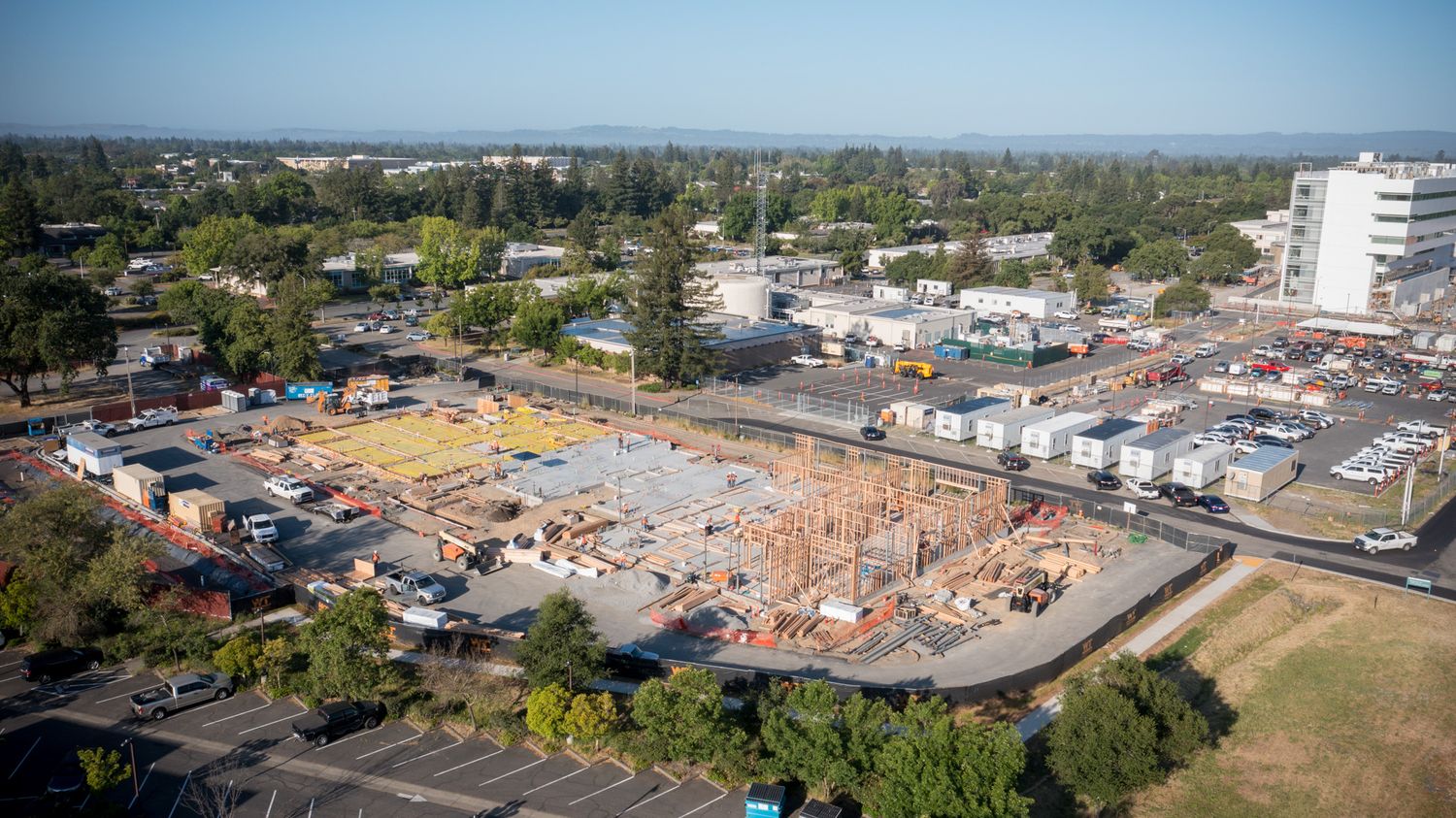 Aerial view of a construction site with orange foundation work visible among parked cars and surrounding buildings.