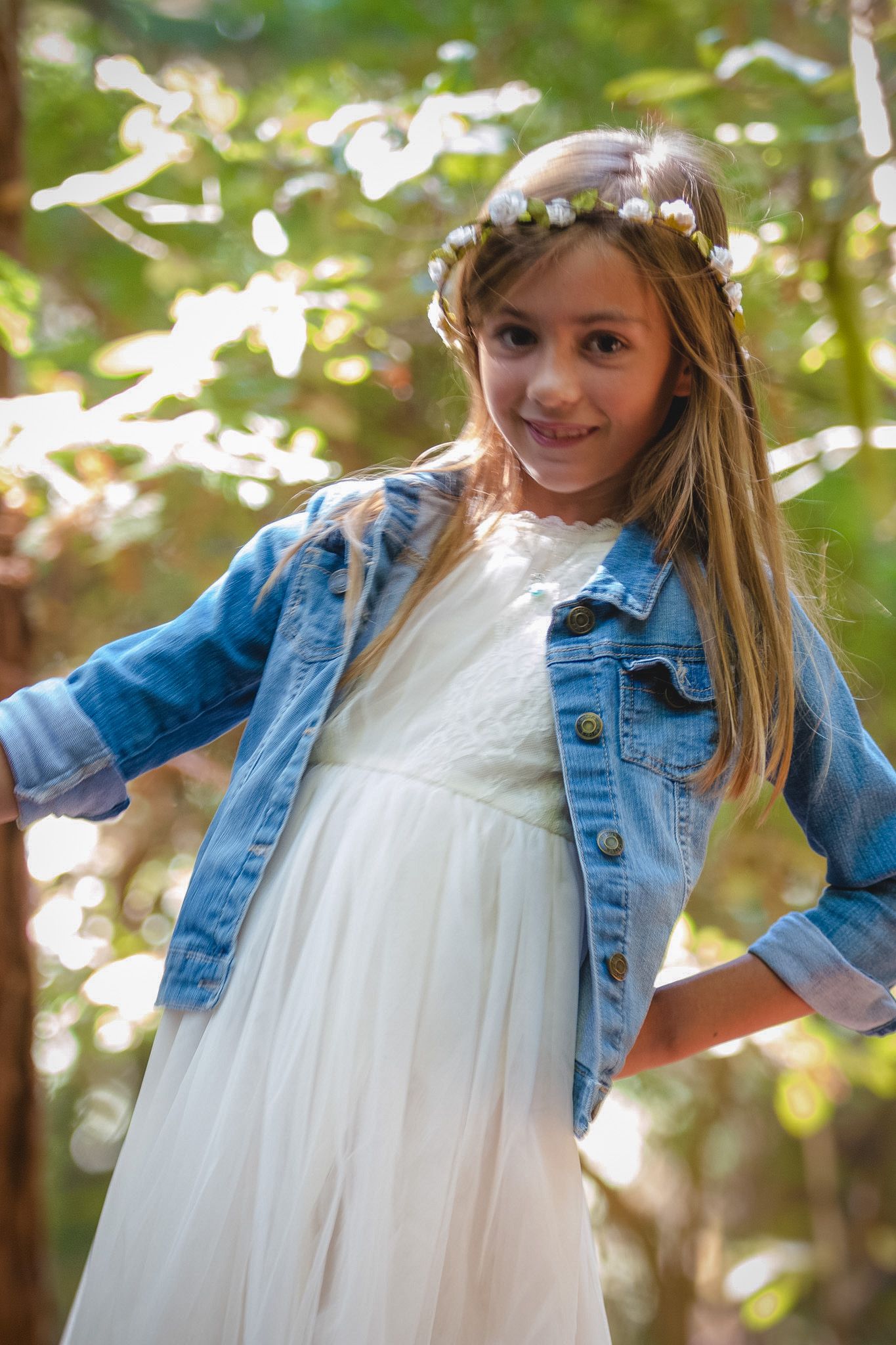 Playful portrait with white dress and denim jacket against a sunlit forest backdrop.