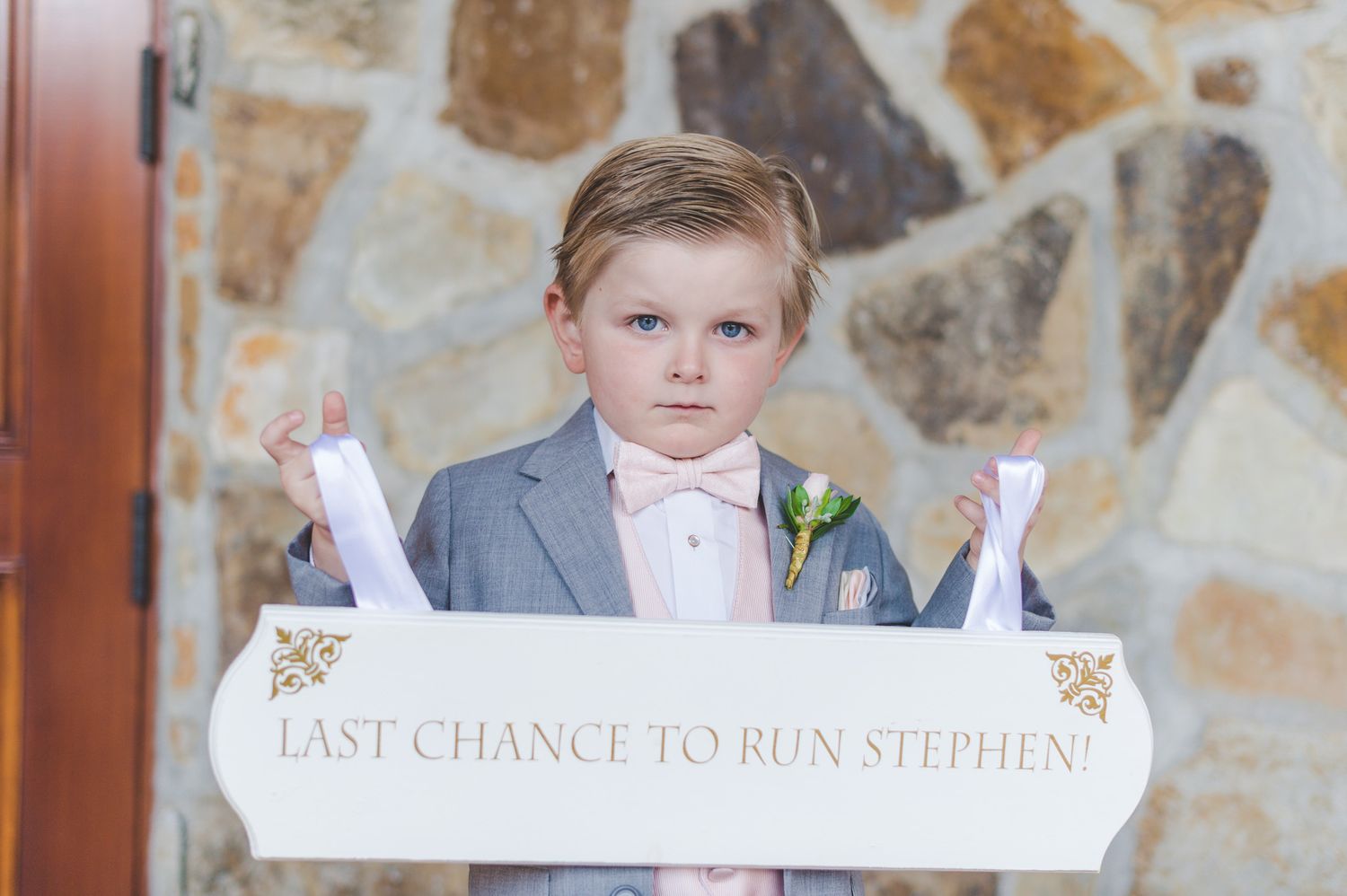 A young attendant in a gray suit holds a white wedding sign against a stone wall backdrop.