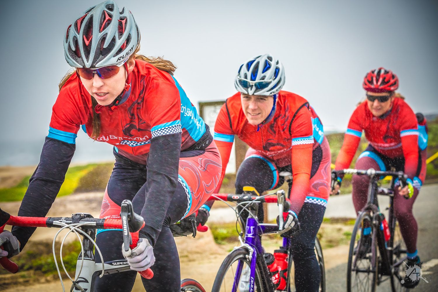 Three cyclists in matching red and blue jerseys ride together during a group training session on a cloudy day.