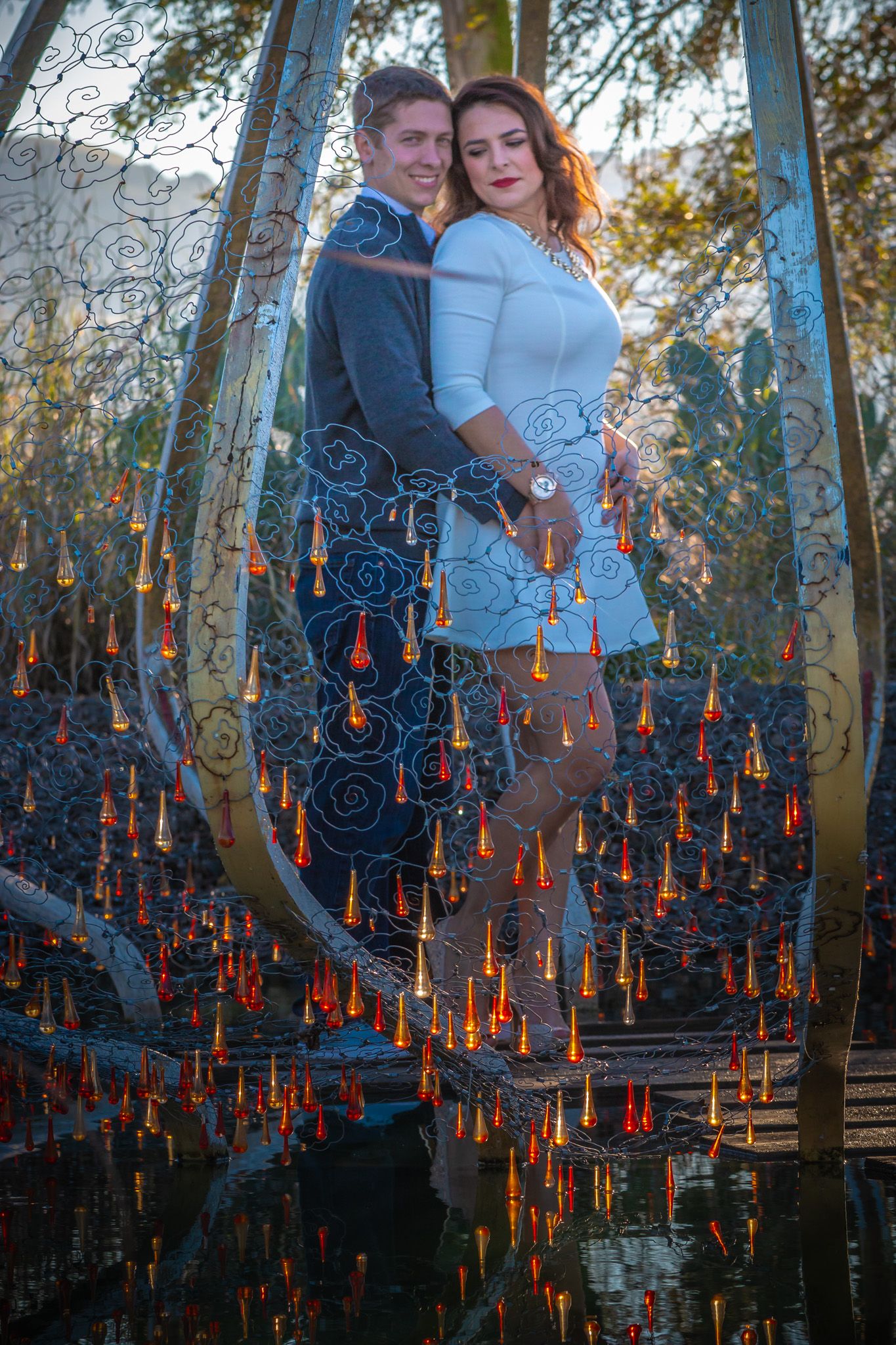 Romantic couple pose on bridge under glowing twilight with hanging orange lights and decorative metal scrollwork.