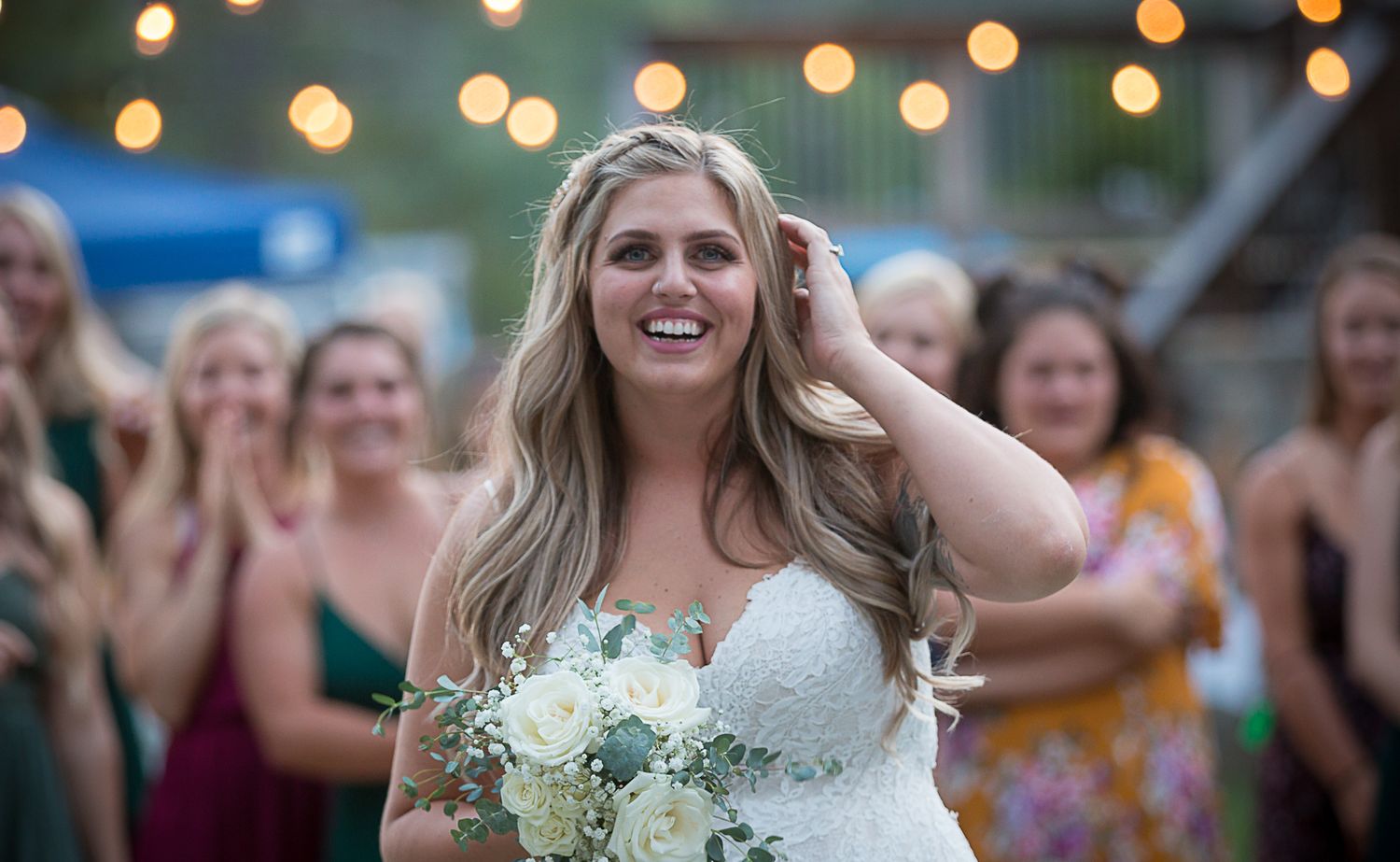 A bride in a white dress holds a bouquet while smiling with glowing string lights in the background.
