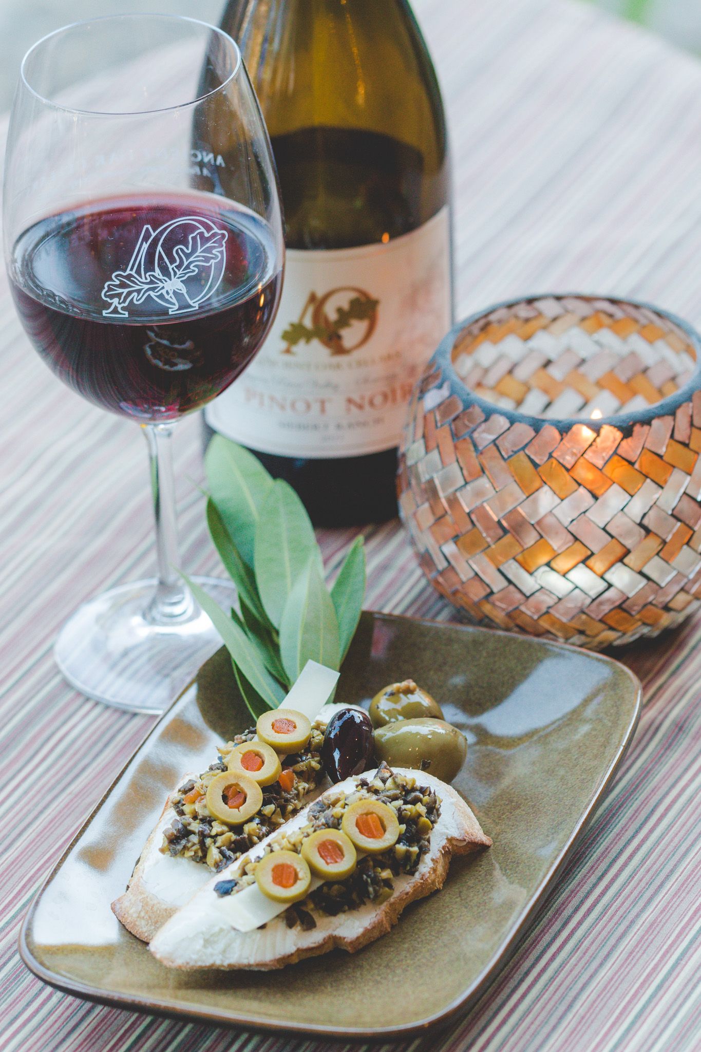 Glass of red wine with Pinot Noir bottle and appetizer plate featuring olives on toast.