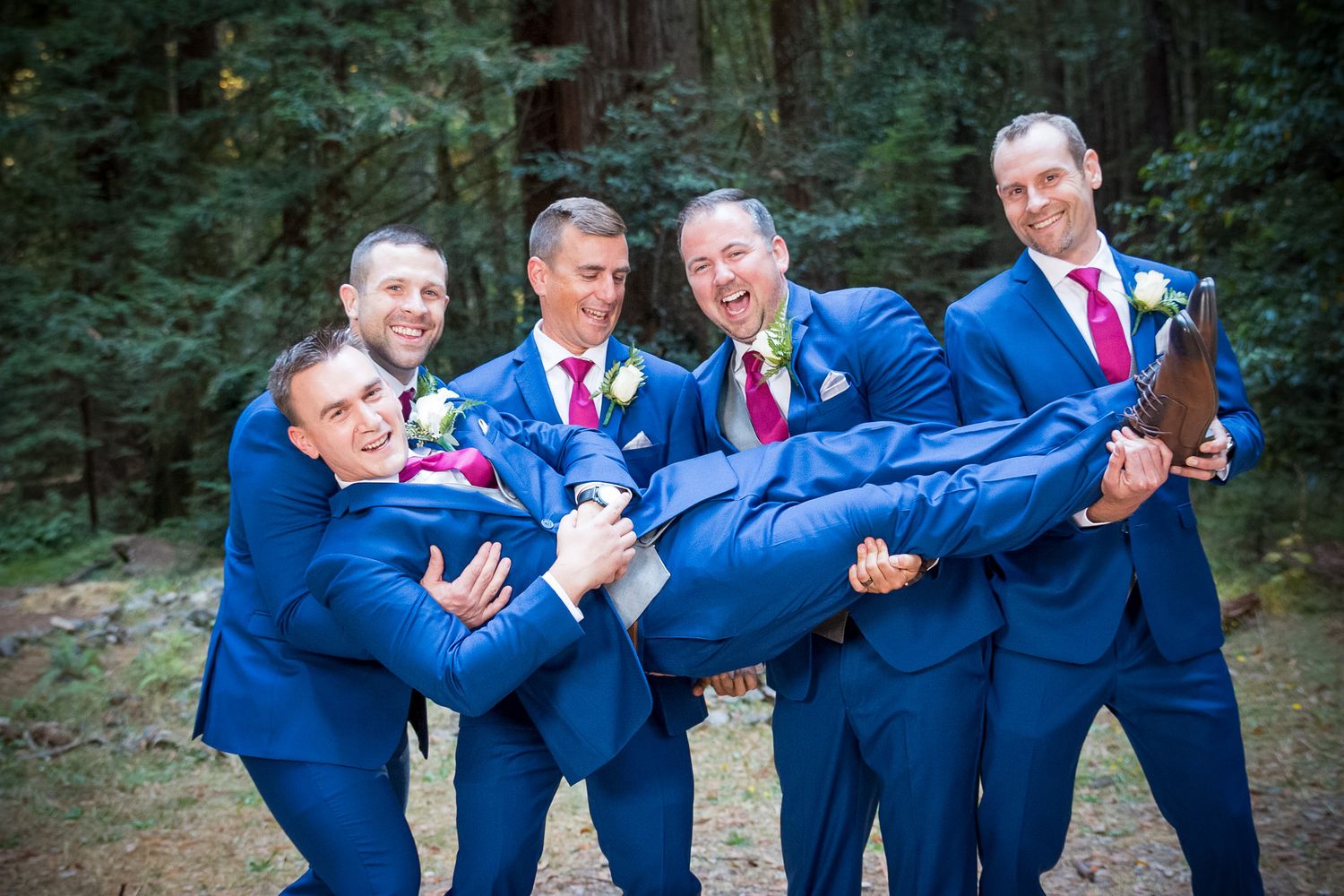 Groomsmen in blue suits playfully carrying groom in outdoor wedding photo.