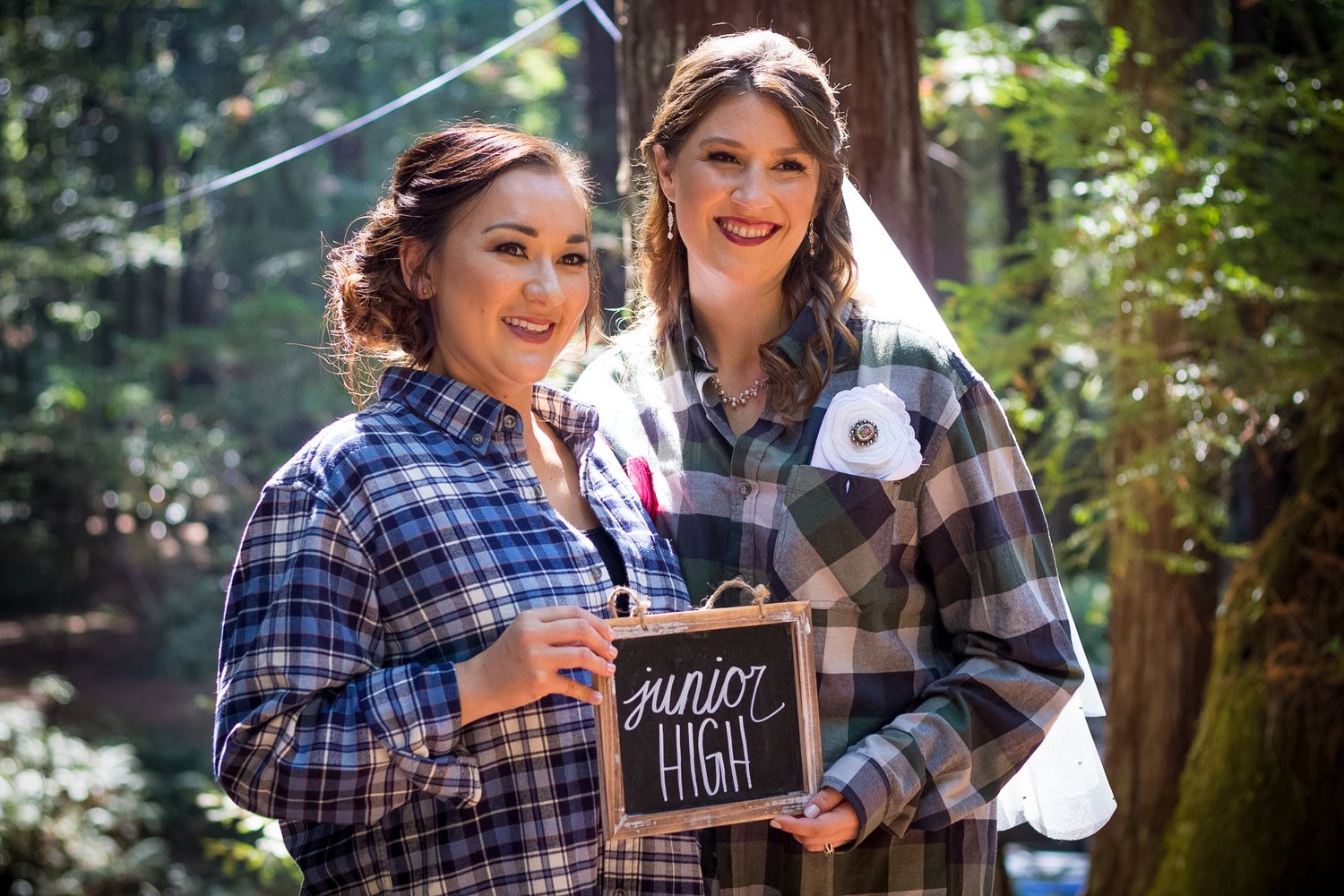 Two people in plaid shirts hold up a small chalkboard sign in a forest setting.