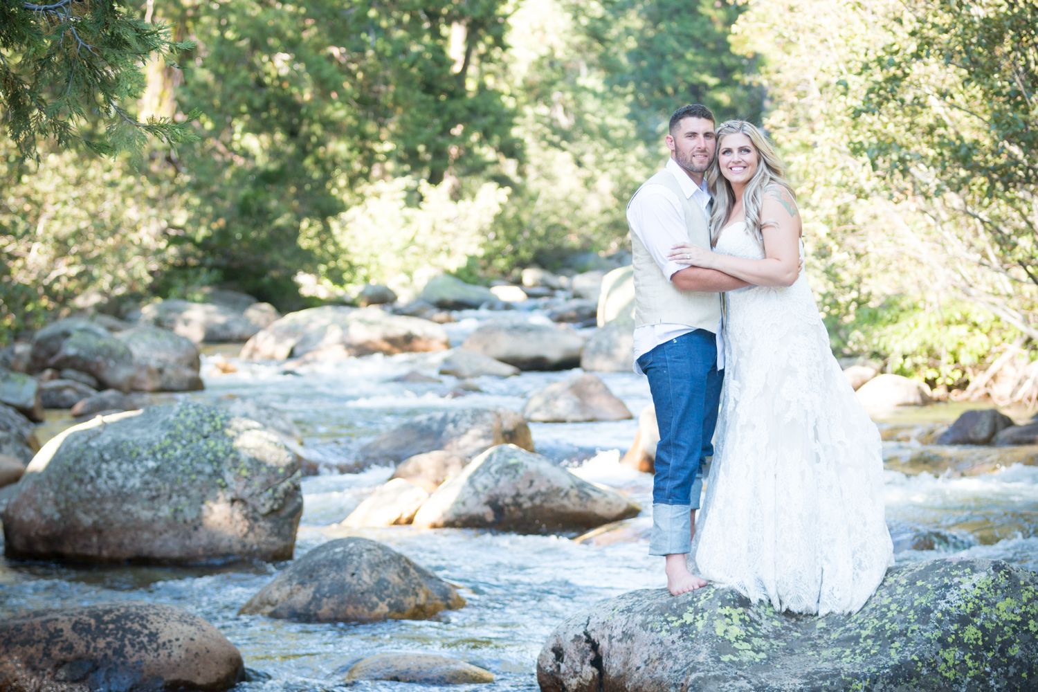 Couple poses on rocky riverbank in wedding attire surrounded by nature.