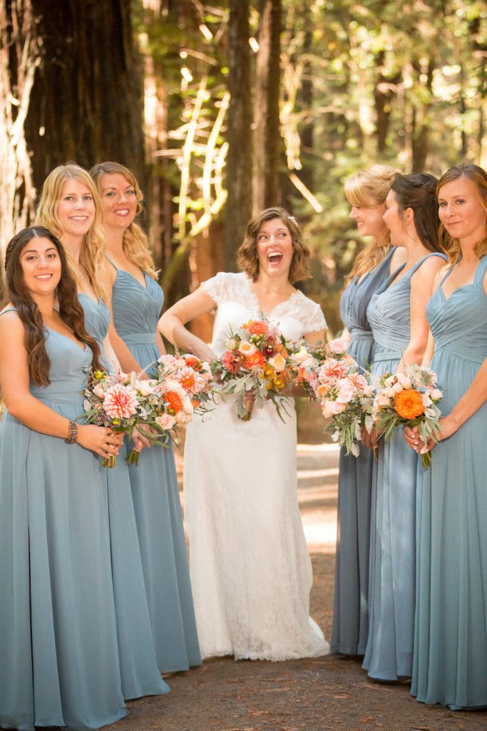Bride and bridesmaids share candid laughter while holding vibrant orange and pink bouquets.