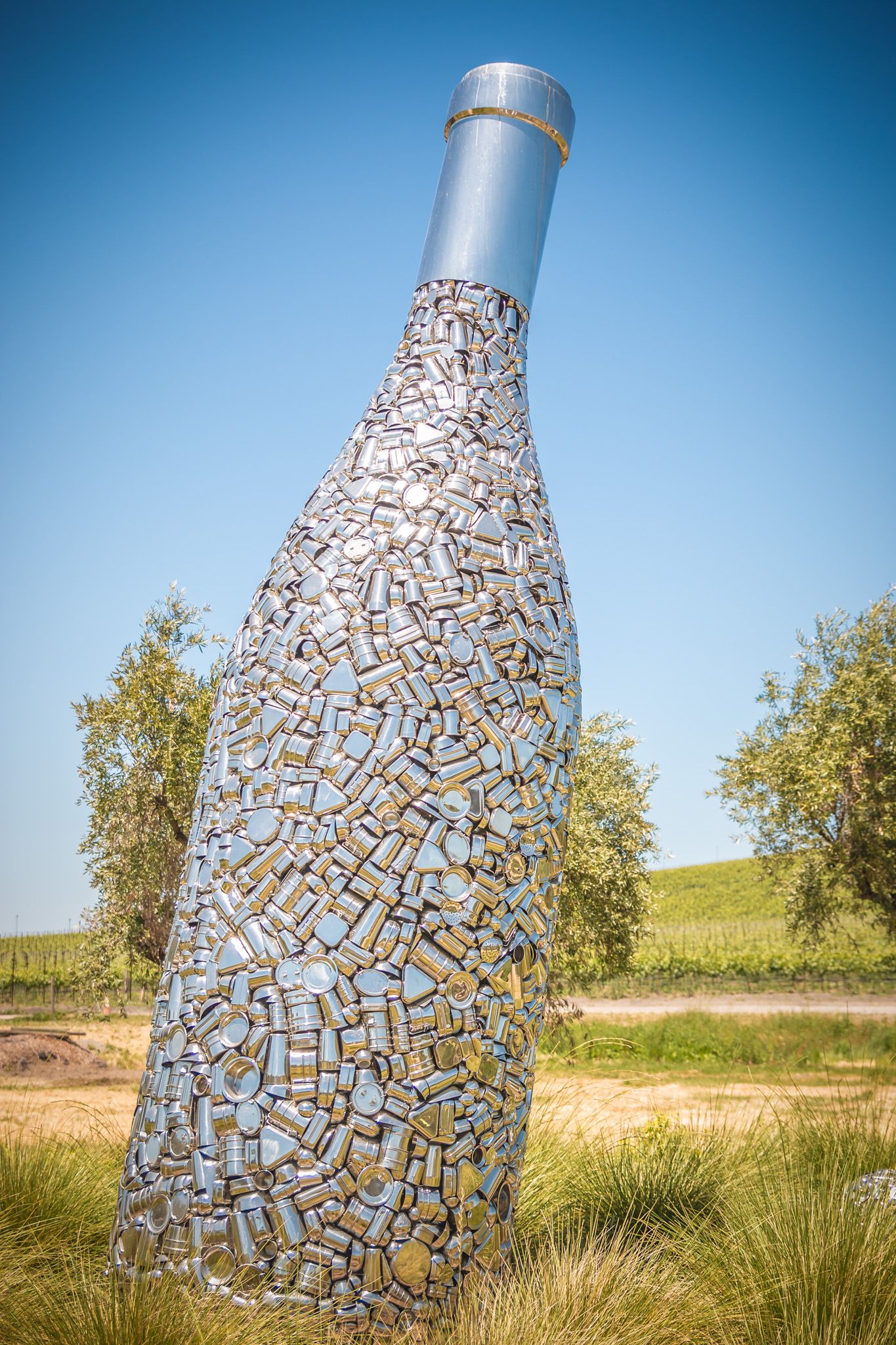 Giant decorative wine bottle sculpture made from hundreds of cork pieces stands in a field.