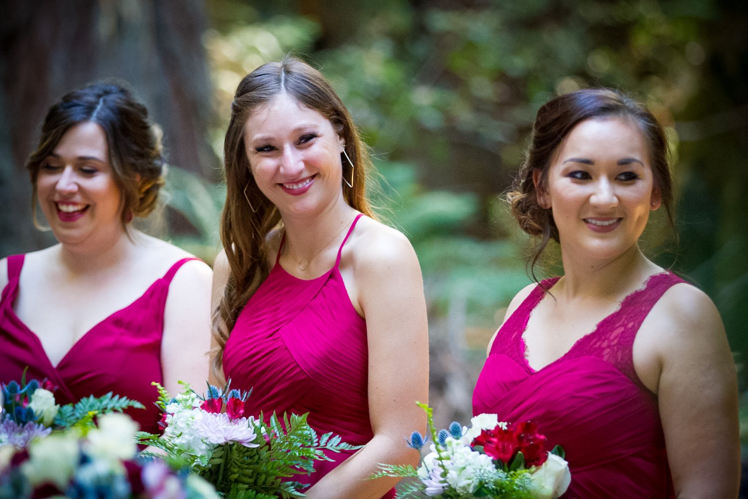 Three bridesmaids in burgundy dresses hold bouquets while smiling outdoors.