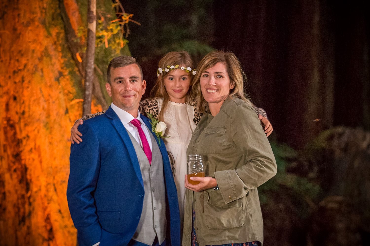 Wedding guests pose together against illuminated forest backdrop at evening reception.