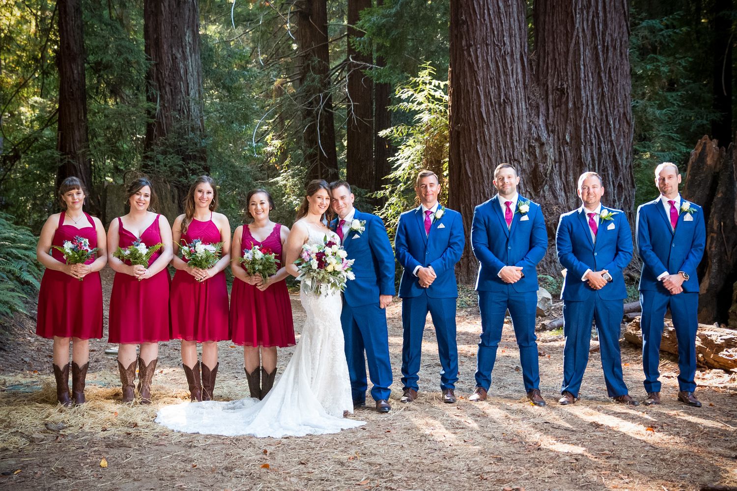 Wedding party in red dresses and blue suits poses among tall redwood trees in a forest setting.