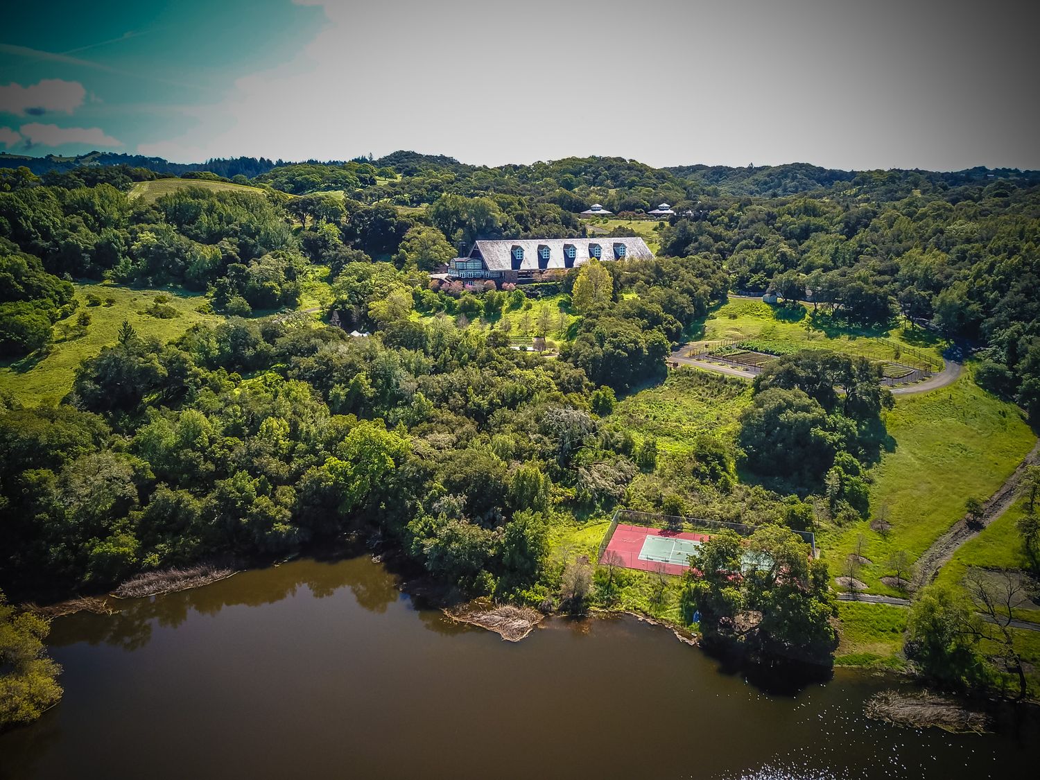 Aerial view of a white mansion with tennis court nestled among green forest near a lake.