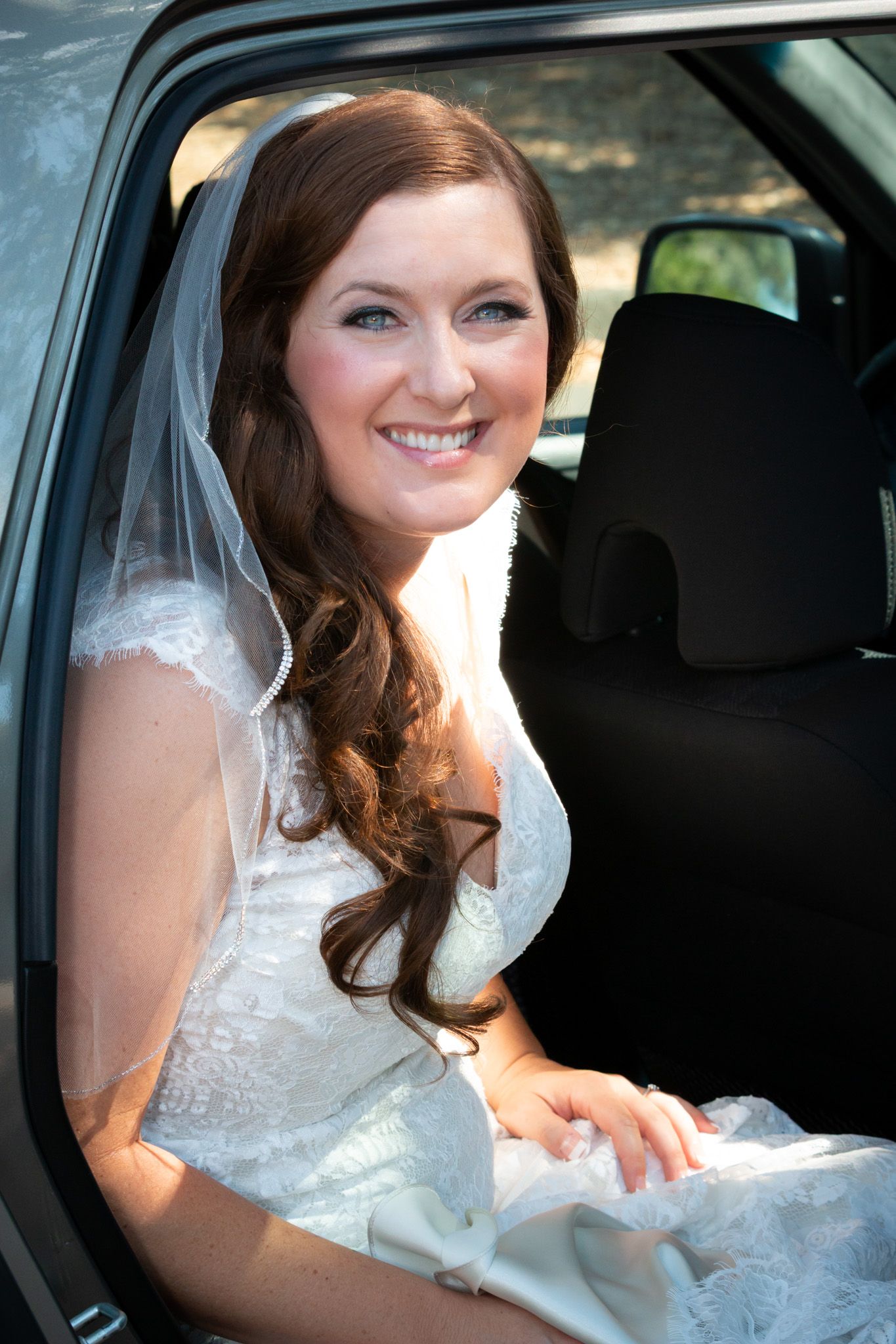 A radiant bride in a white lace wedding dress sits elegantly in a car.