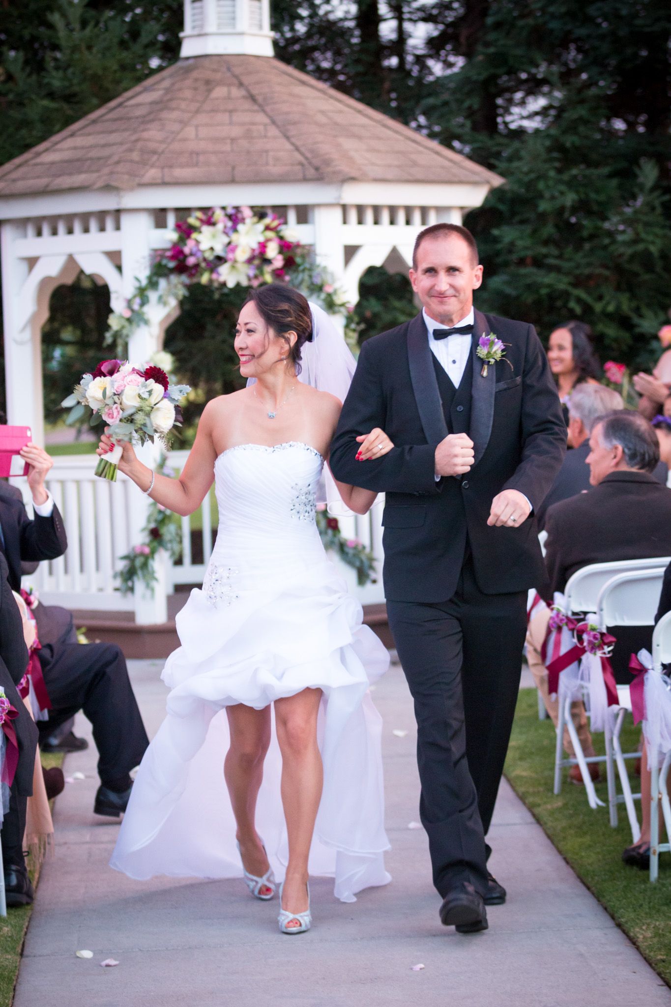 Cheerful newlyweds walk down the aisle after ceremony at outdoor gazebo decorated with pink and white floral arrangements.