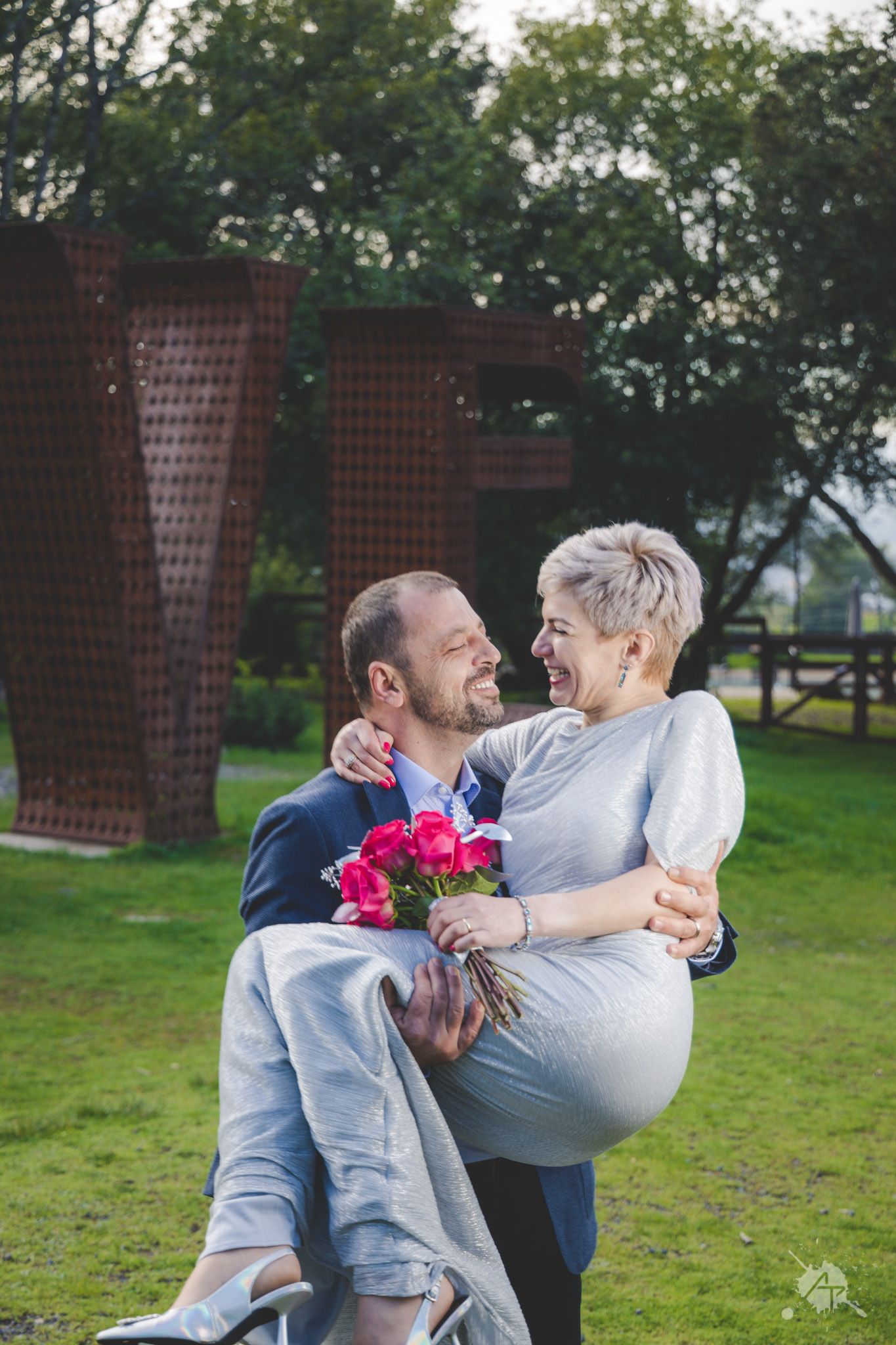 A joyful couple embraces in a park, holding pink flowers with large rustic metal sculptures in the background.