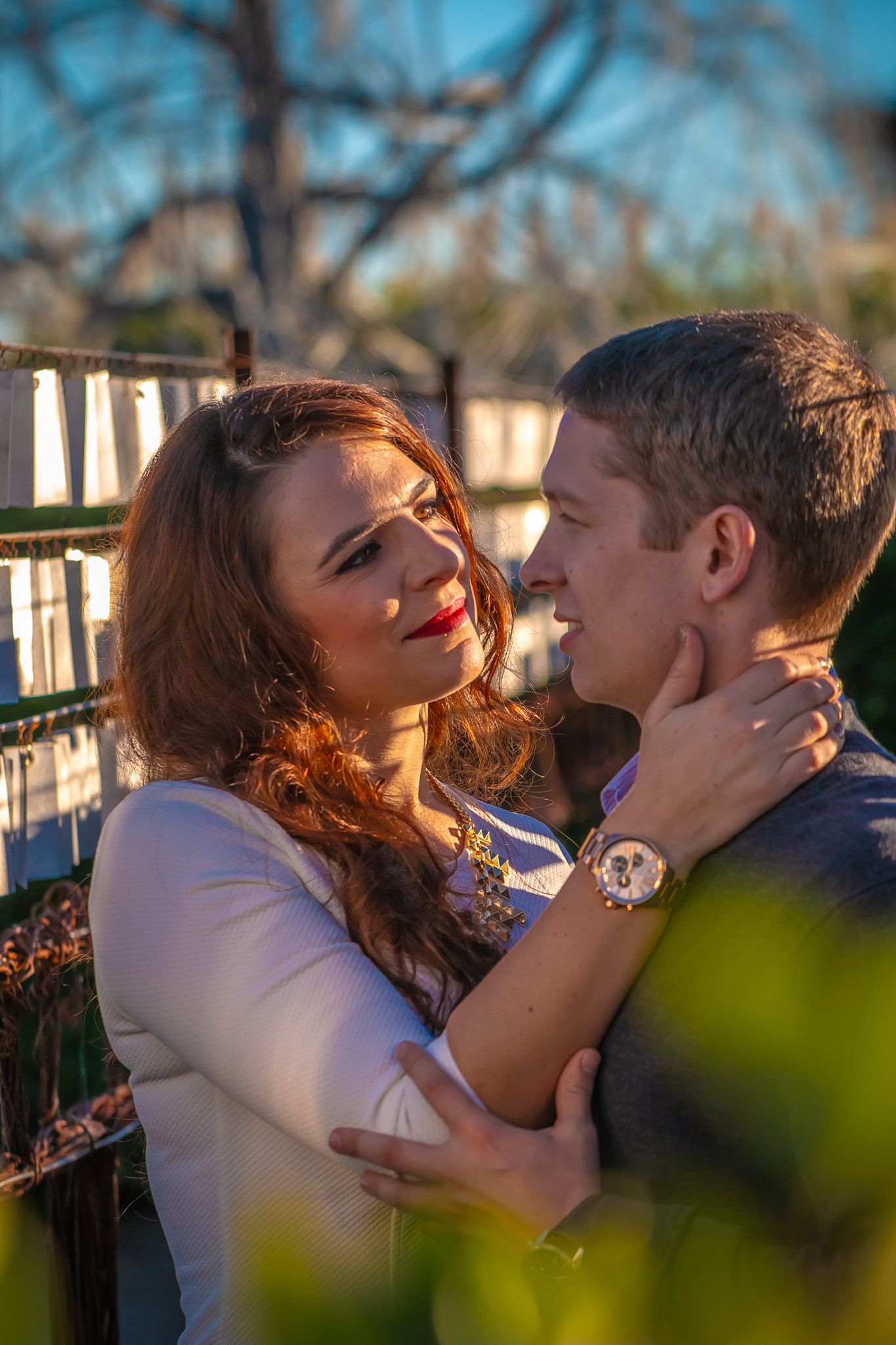 Golden hour portrait of couple in loving embrace near white fence with bokeh effect.