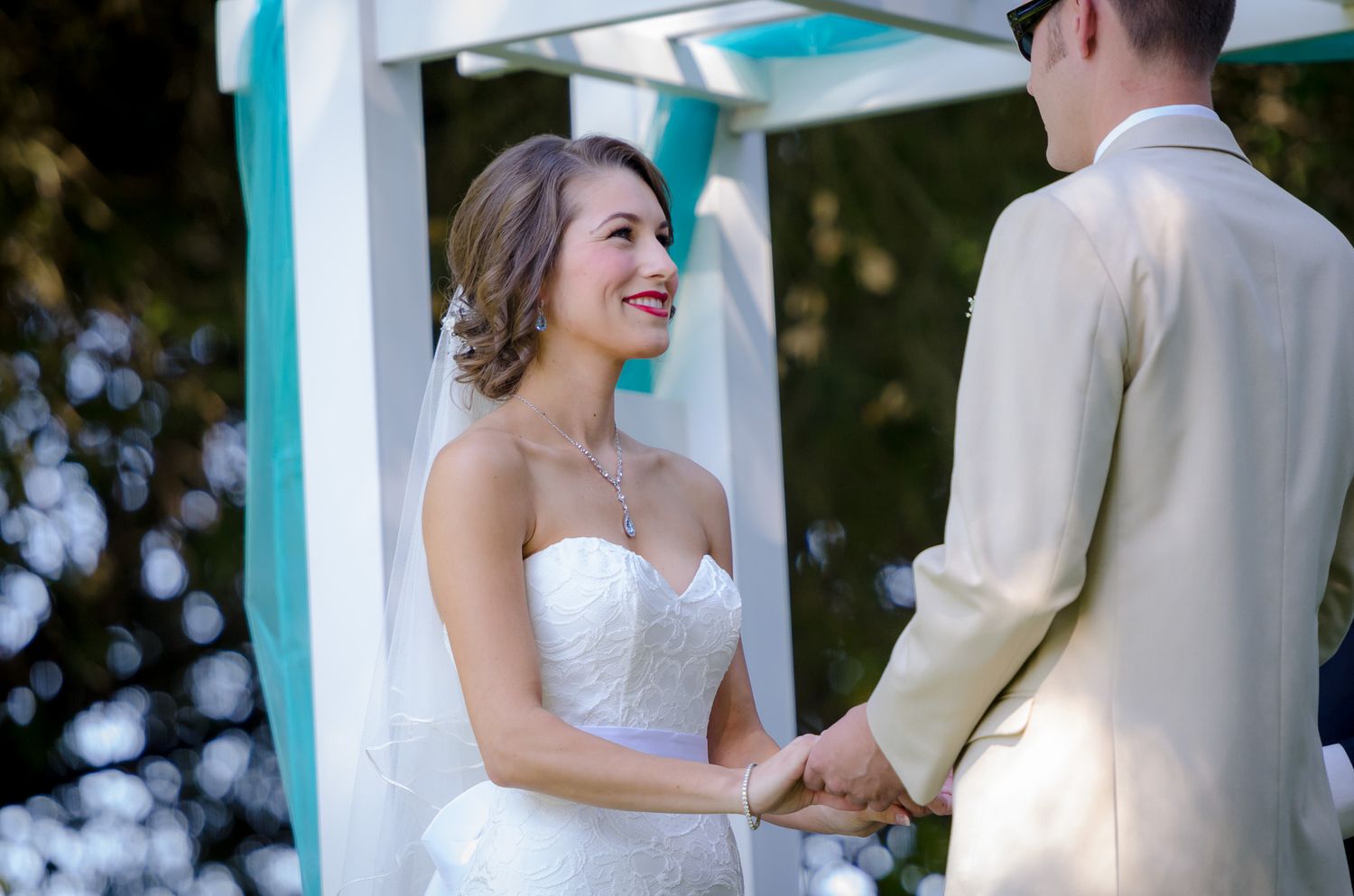 Happy couple holds hands during romantic outdoor wedding ceremony under white gazebo.