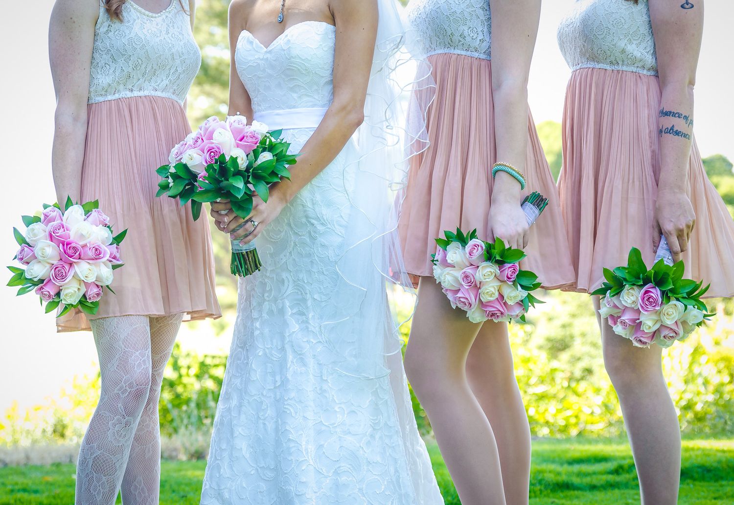 Bridesmaids in pink and mint dresses stand beside bride in white lace gown holding pink floral bouquets outdoors.