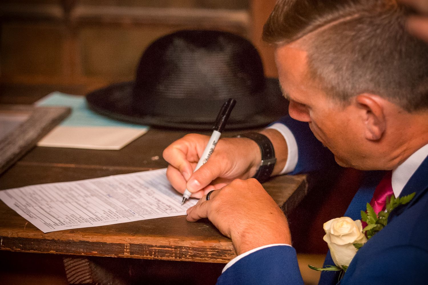 A person in a blue suit signing documents with a black fedora on the desk.