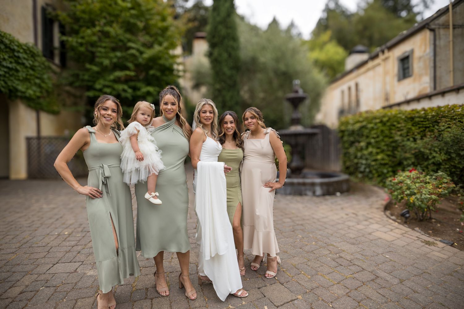 Wedding party in sage green dresses poses together on brick courtyard with bride in white gown.
