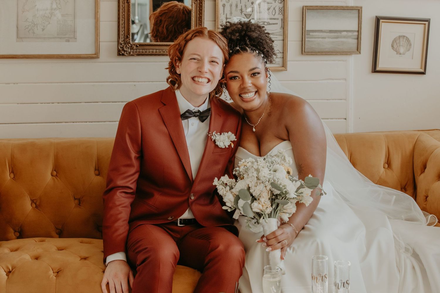 A happy newlywed couple posing on vintage yellow couch, wearing white gown and rust-colored suit.