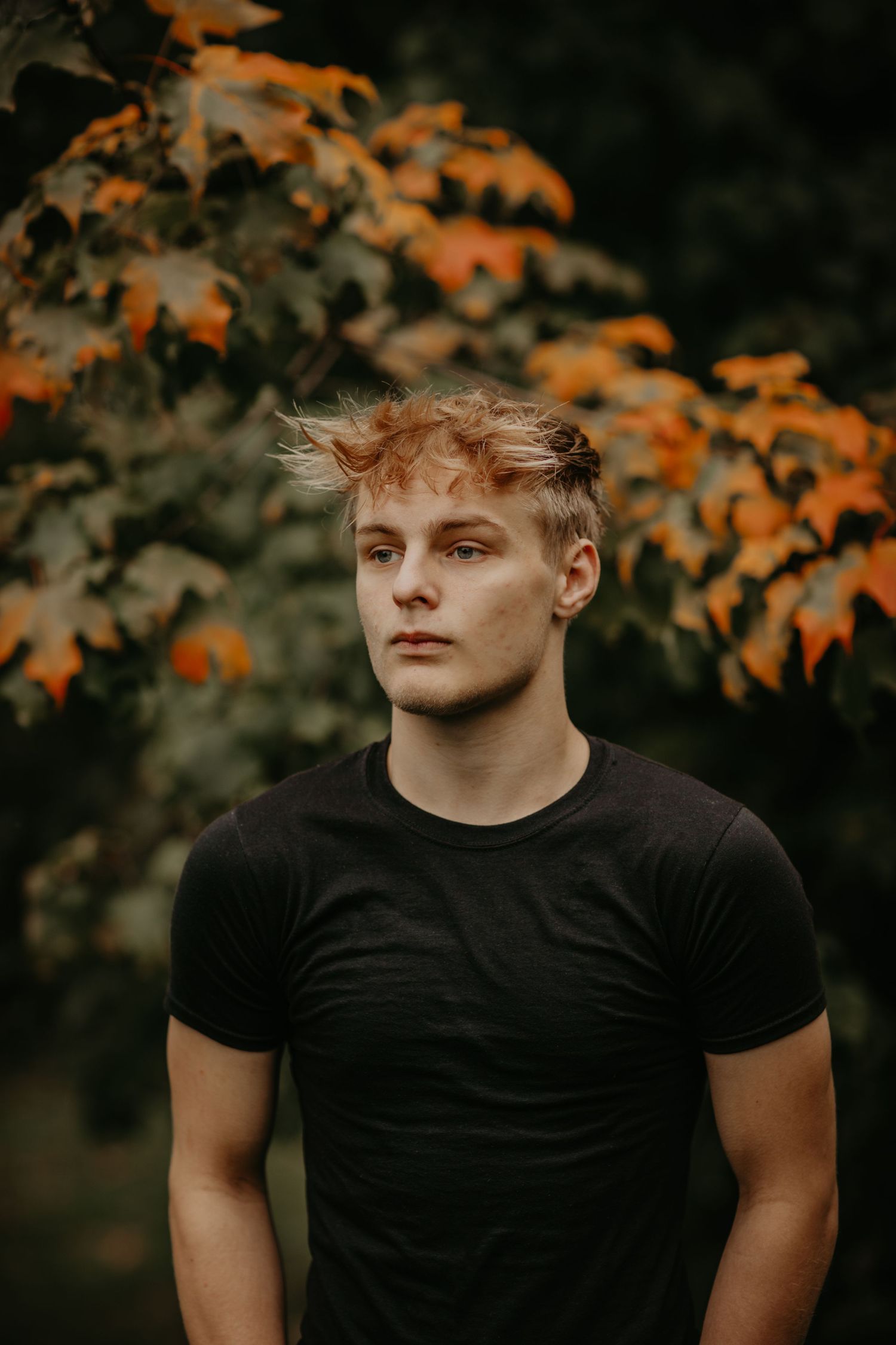 Portrait photo in black t-shirt against autumn foliage backdrop with orange leaves and moody lighting.