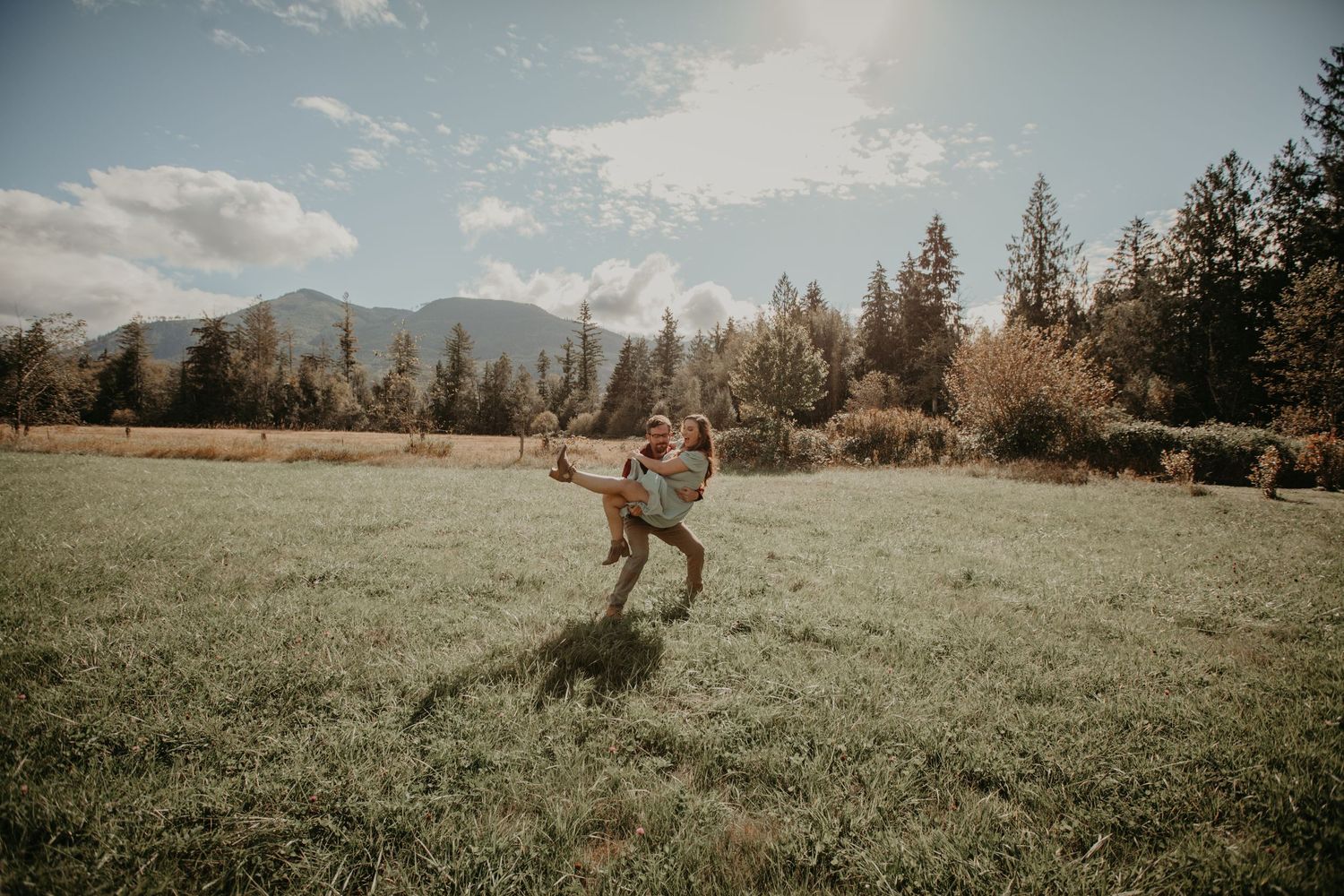 A person dances in a grassy meadow with mountains and evergreen trees visible in the background under a cloudy sky.