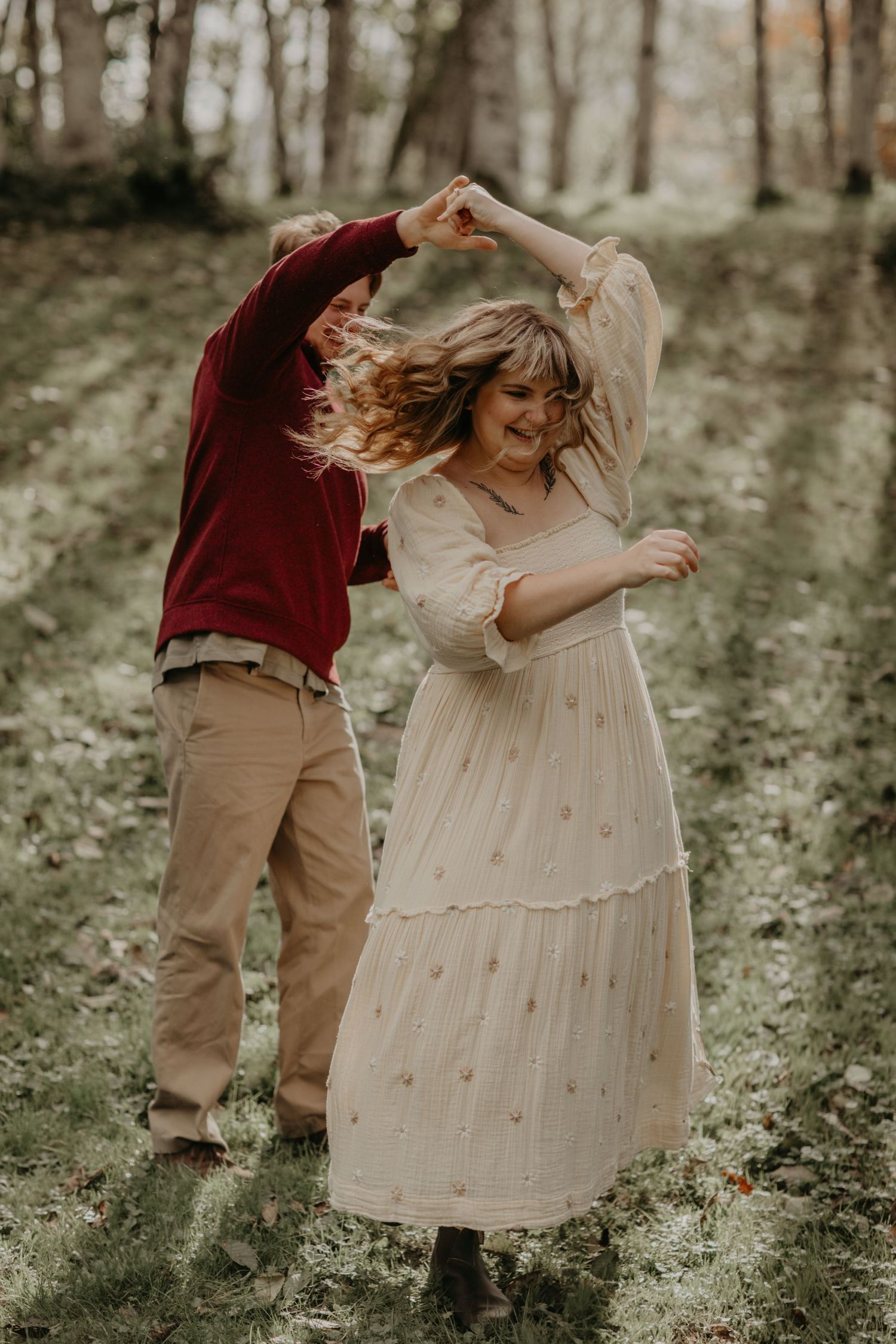 A couple dances joyfully together in a wooded setting, with the woman wearing a light cream dress and partner in burgundy.