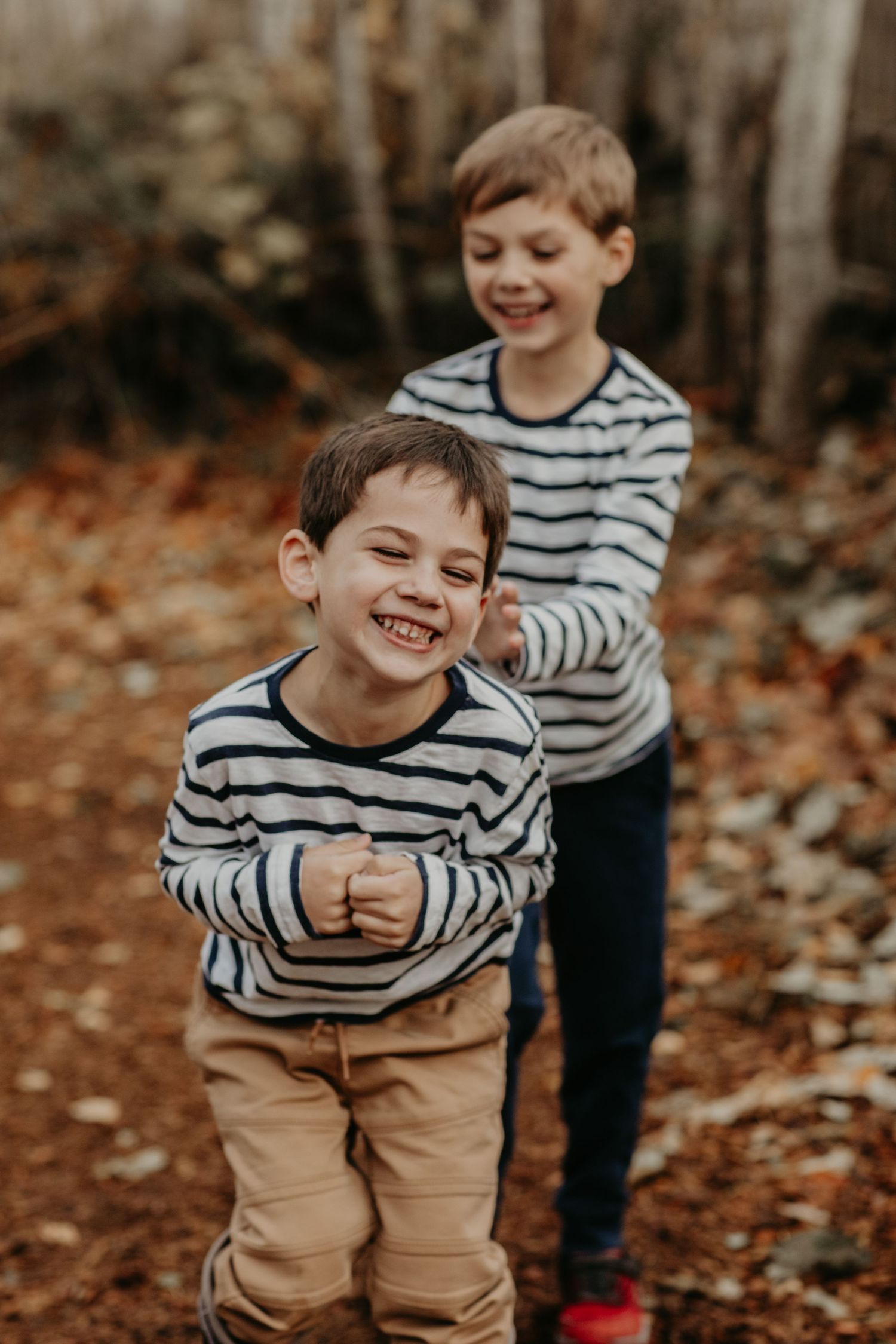 Two children in matching striped sweaters laugh together among fallen autumn leaves in the woods.