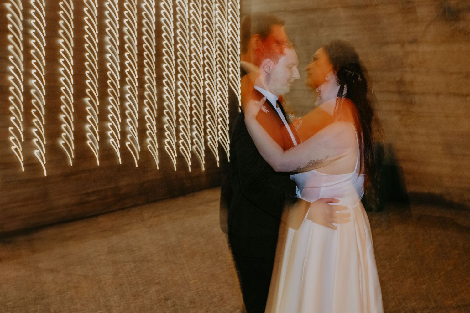 A couple dances together at their wedding reception under vintage-style lighting in a rustic wooden room.
