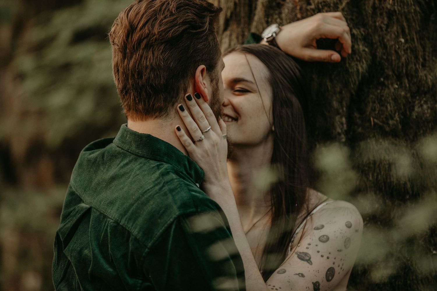 Two people share an intimate romantic moment against a tree in moody natural lighting.